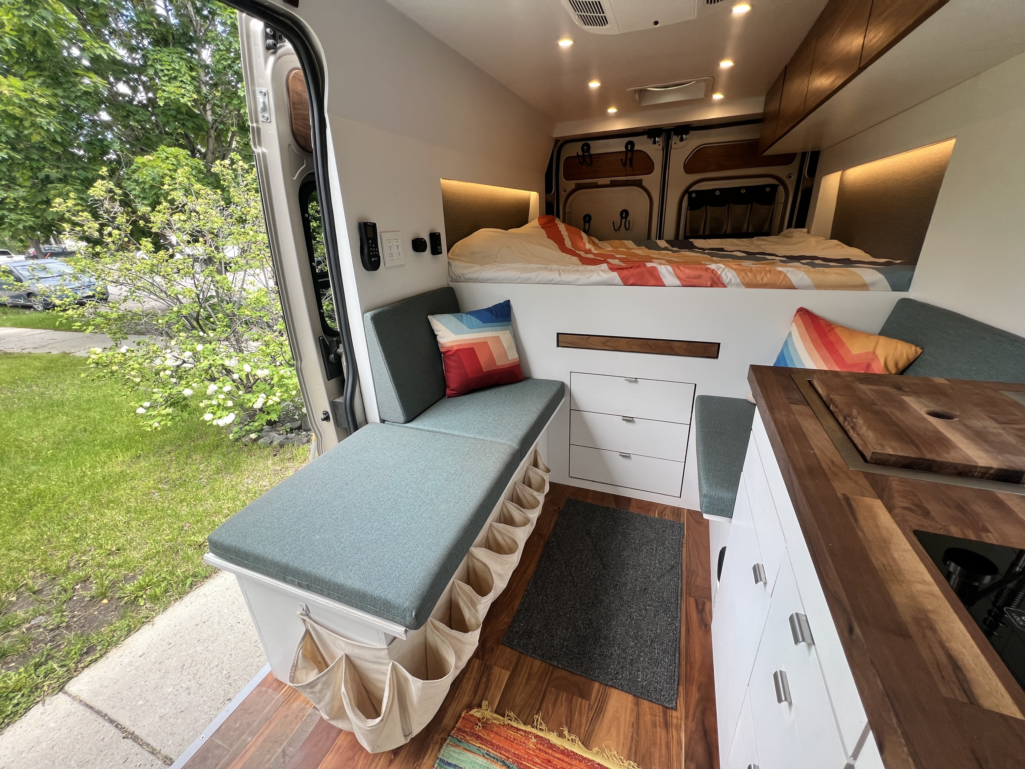 Cozy RV rental interior featuring an elevated bed, compact kitchenette, bench seating with storage, and green outdoor views.. Dodge Ram Promaster 2500 2021