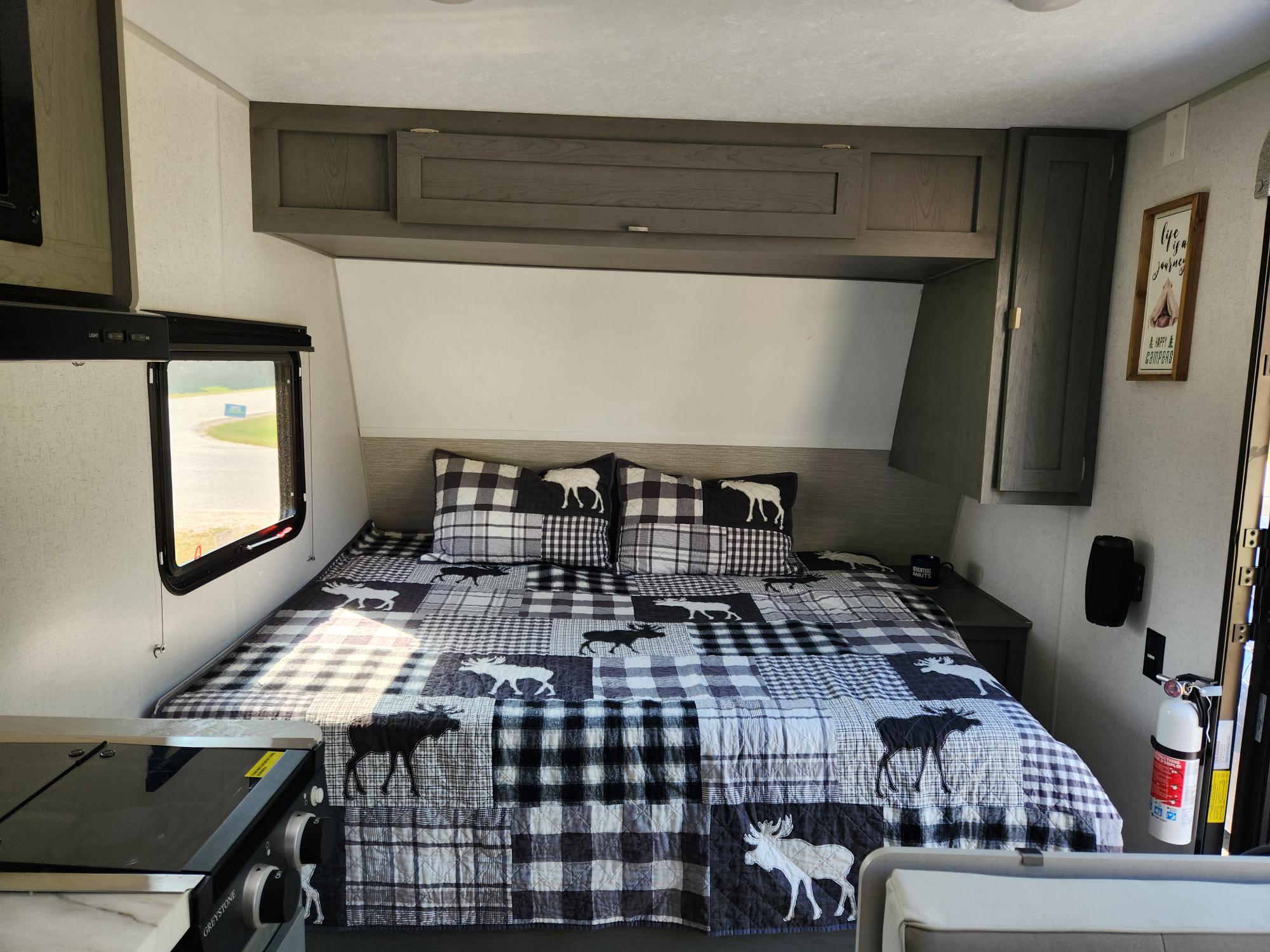 Cozy RV rental bedroom with a moose-themed quilt, overhead storage, and a partial kitchenette, perfect for travel.. Ozark Ozark Trailer 2023