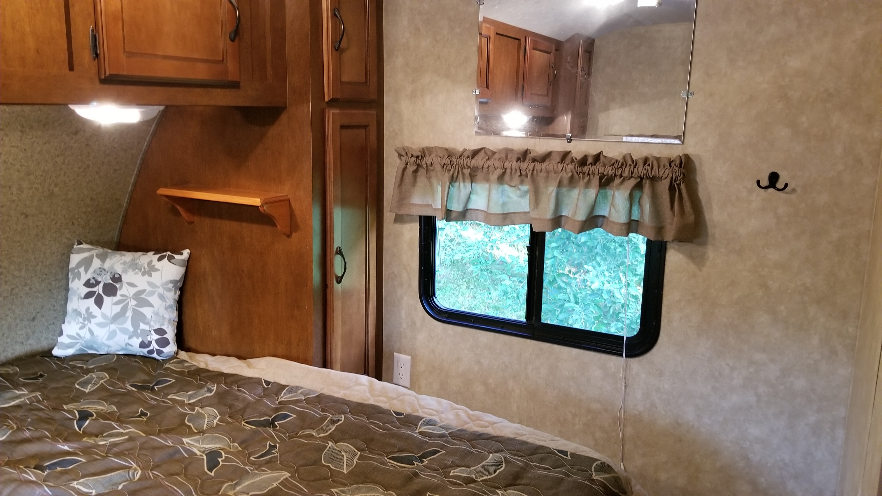 An RV rental's bedroom area shows a patterned bed, overhead cabinets, a small shelf, and a window with outside greenery.. Forest River Coachmen Catalina 2013