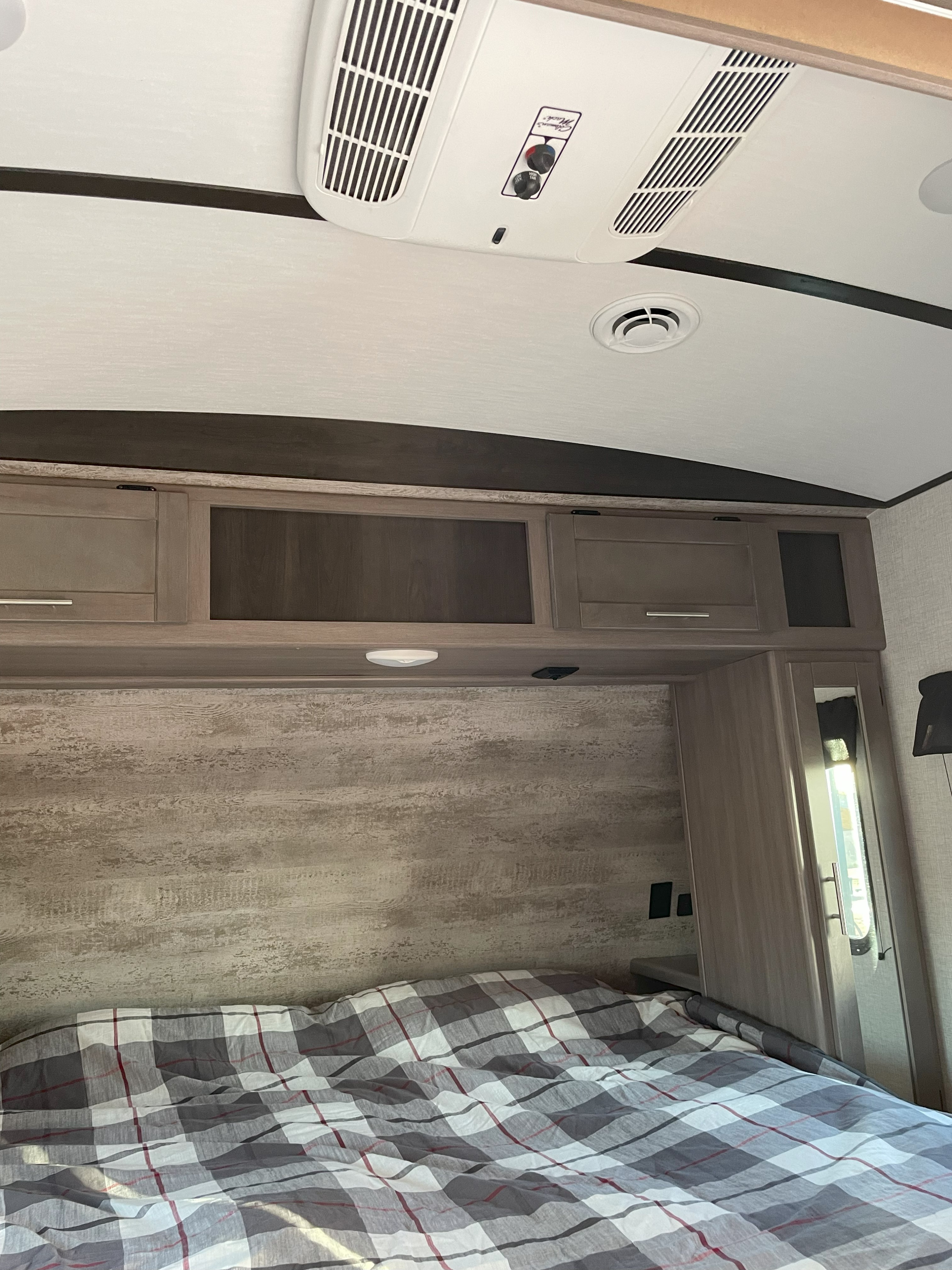 RV rental interior: Plaid comforter on bed, textured headboard, overhead cabinets, and ceiling air conditioner.. Sunset Park RV 272BH 2022