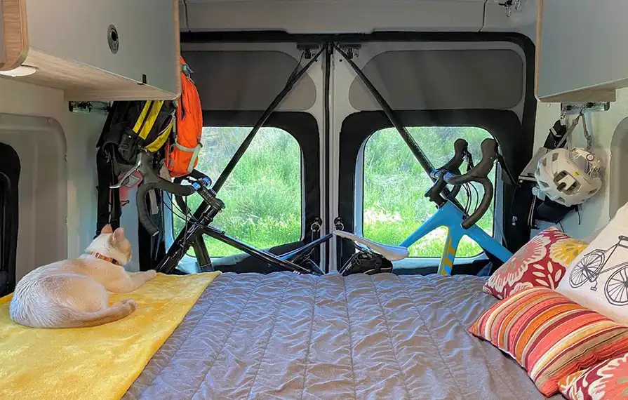 Inside an RV rental: a cozy bed with a cat, clever bike storage, & hanging gear. Ideal for adventure travel.. Winnebago Solis 2021