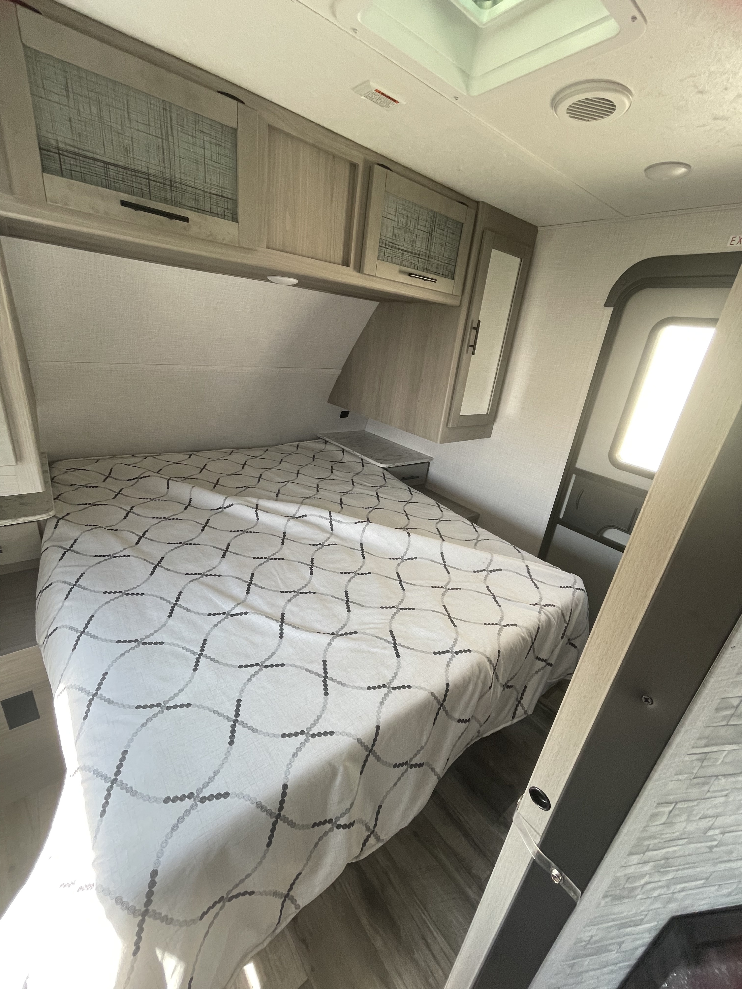This RV rental's bedroom features a bed with patterned cover, light wood cabinets, and bright lighting from the nearby exit.. Dutchmen Coleman Light 2022