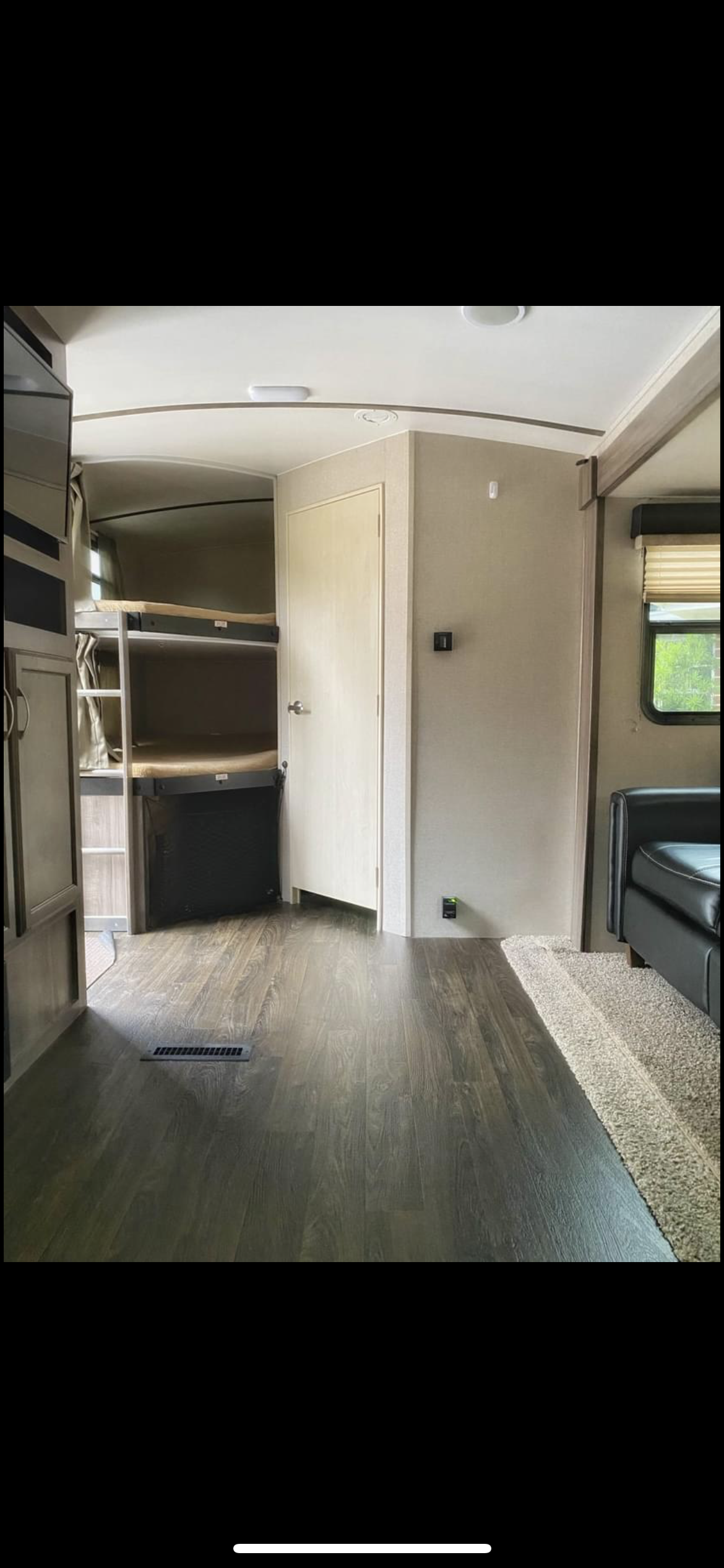 Inside this RV rental, you see dark wood-look flooring leading to bunk beds and a closed door. A beige rug borders a black couch on the right.. Minnie Minnie Plus 2018