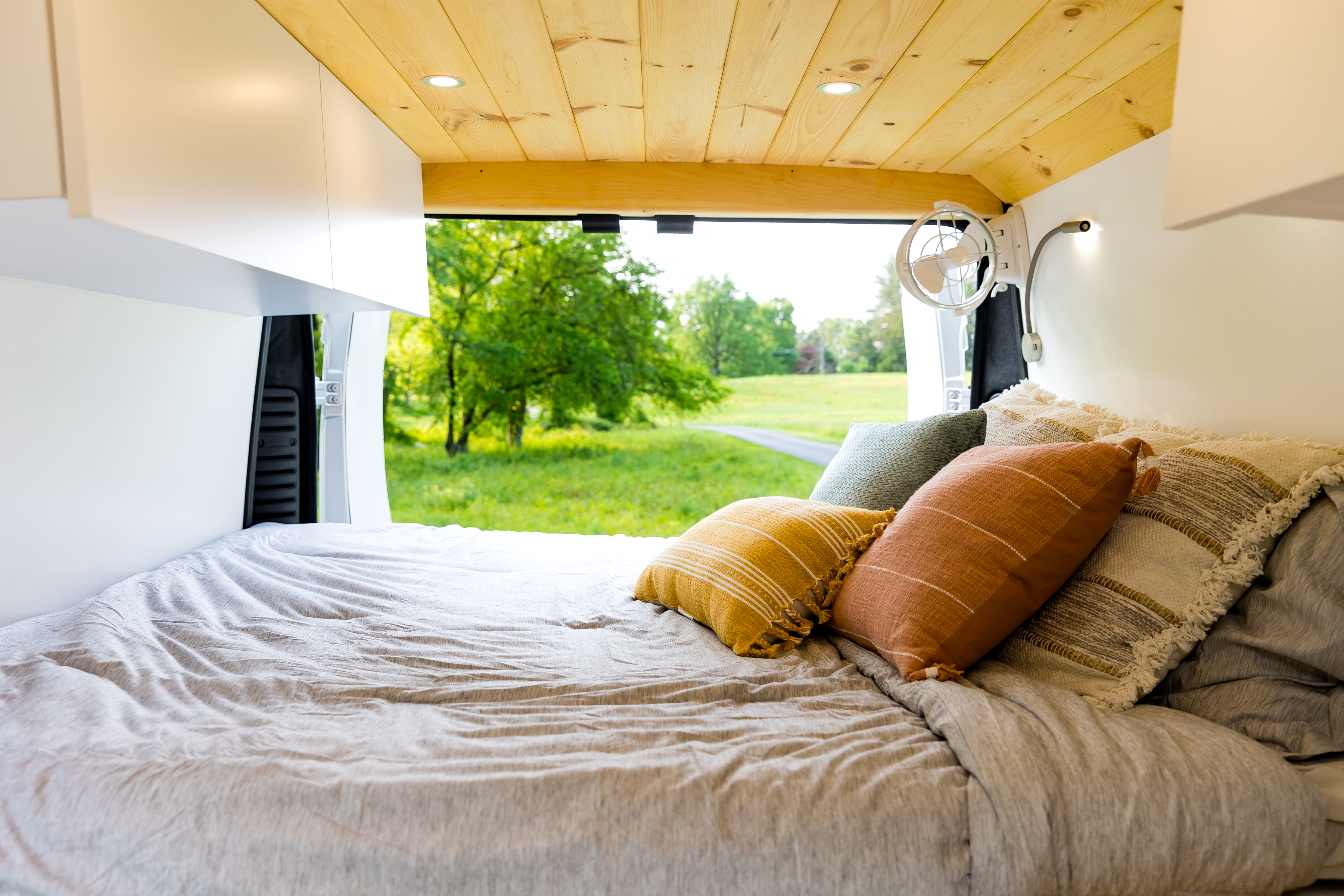 Inside an RV rental: a cozy bed with colorful pillows, overhead storage, and wooden ceiling. Open doors reveal a lush green view.. Dodge 1500 2023