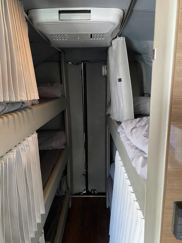 An RV rental interior with multiple bunk beds & privacy curtains. A narrow aisle separates sleeping areas, topped by an AC unit.. Mercedes-Benz Sprinter 2012