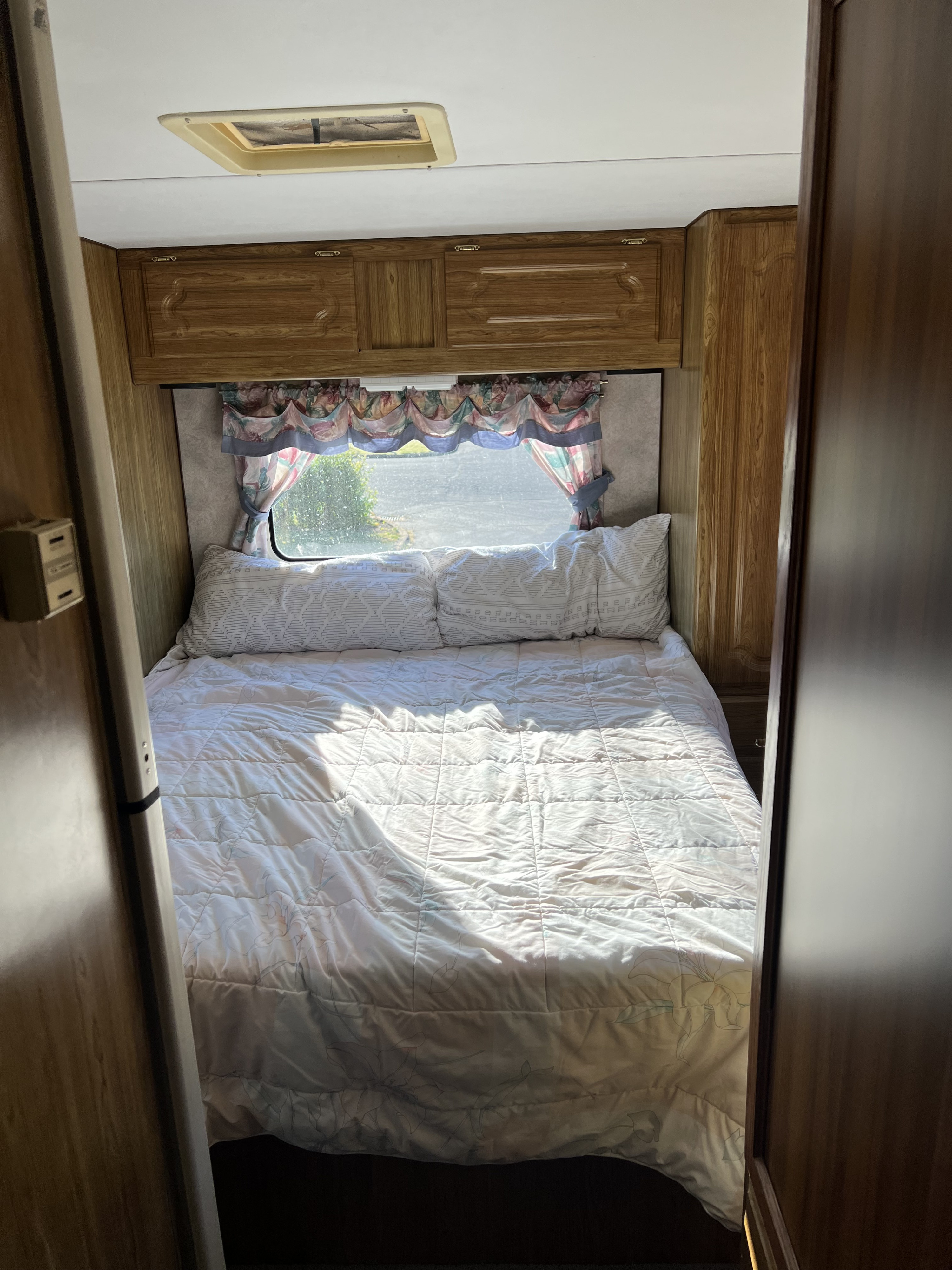 Inside a cozy RV rental bedroom. A neatly made bed with white bedding and pillows, window with floral curtains, and wooden cabinets are visible.. Gulf Stream Conquest 1996