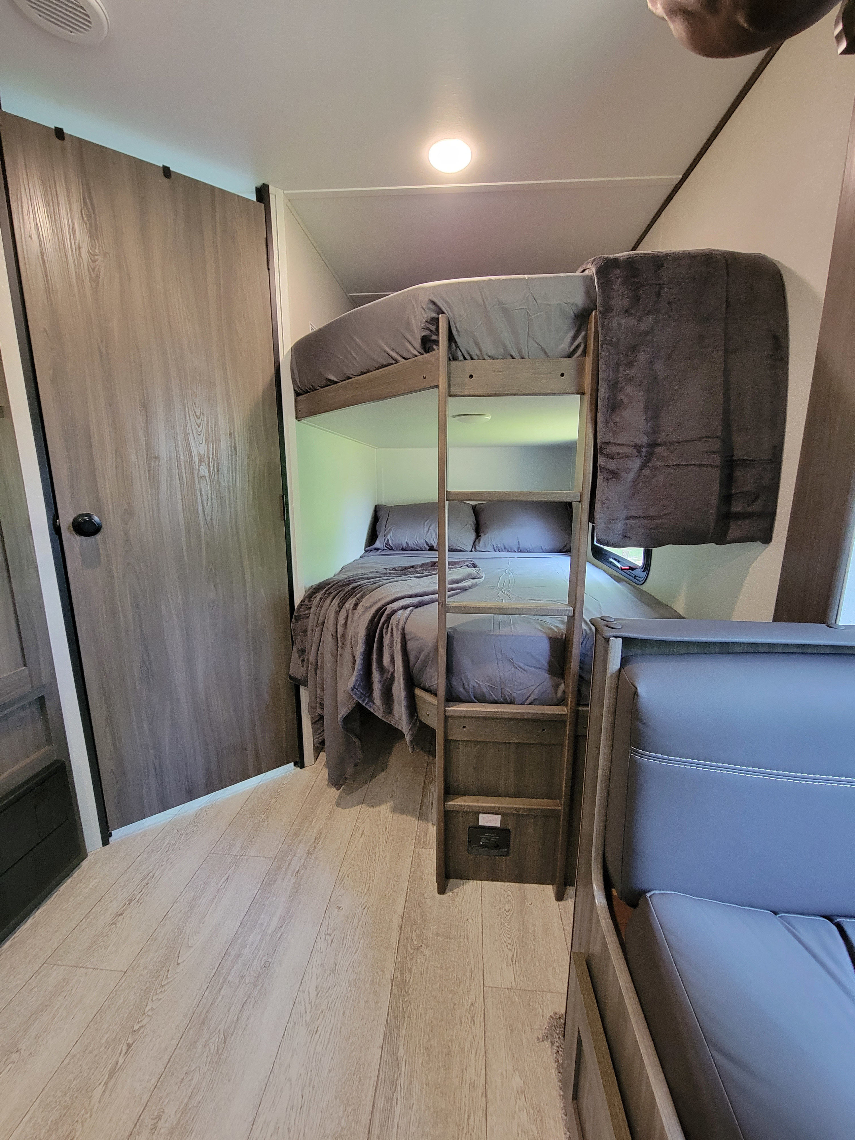 RV rental bunk beds feature gray bedding, a ladder, light wood floor, and a partial view of an upholstered seat.. Gulf Stream Grand River 2021