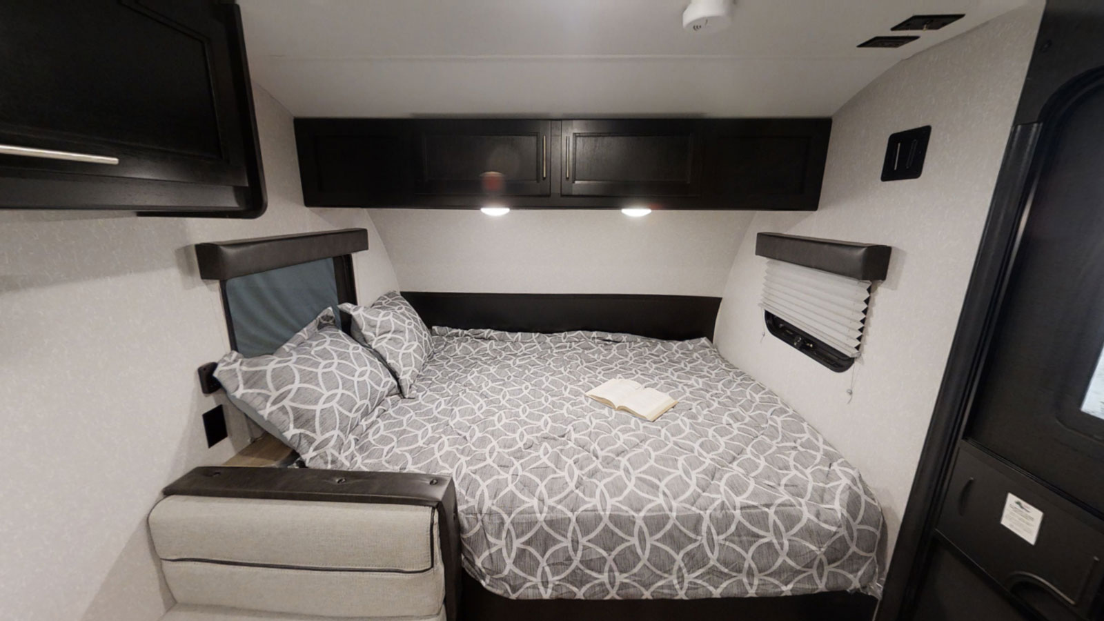 A cozy RV rental bedroom features a grey patterned bed, overhead cabinets, and windows, perfect for comfortable rest.. Forest River Wolf Pup 16BHS 2021