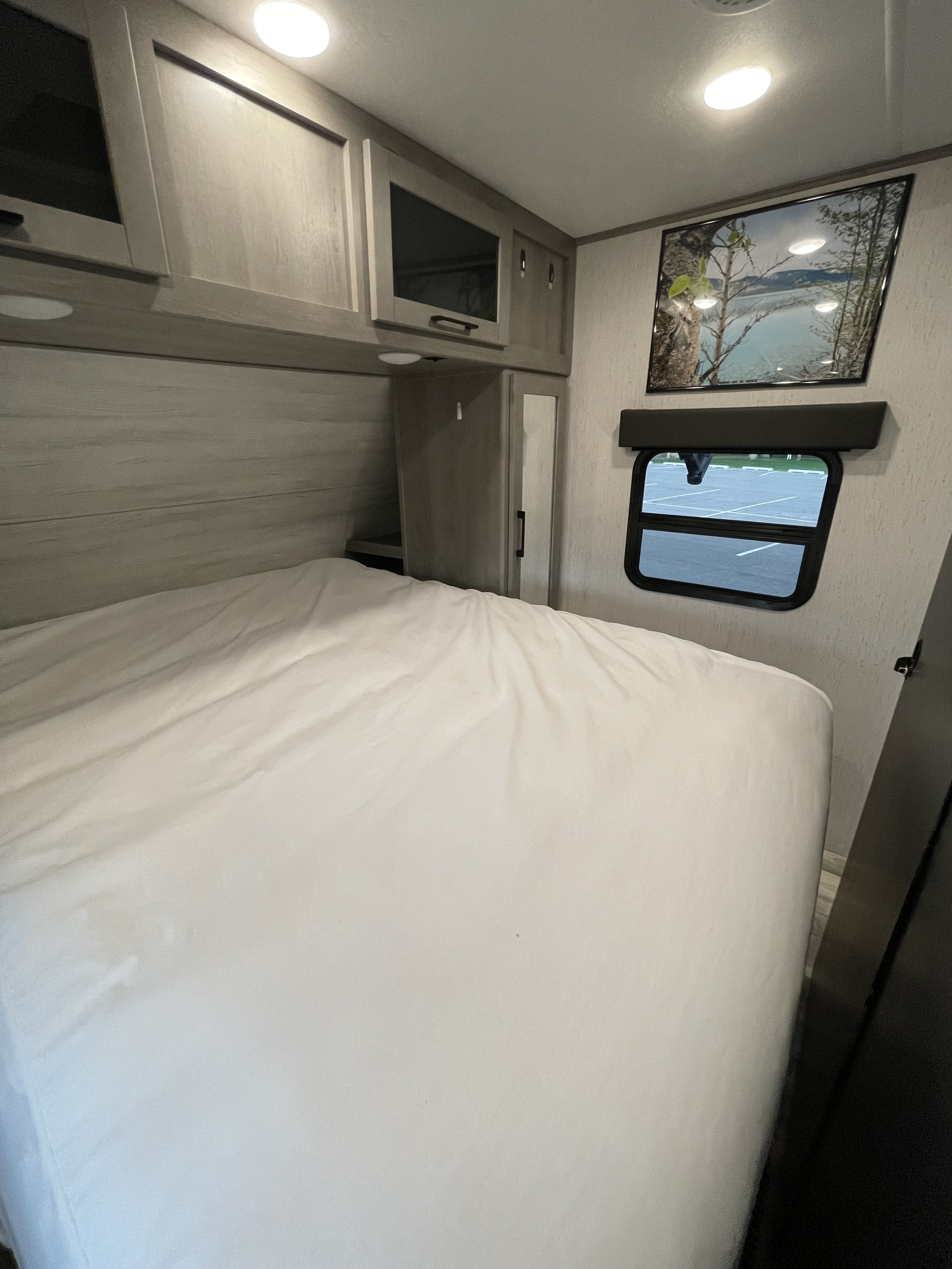 RV rental bedroom: a bright space with overhead cabinets, a white bed, and a picture above a small window.. Dutchmen Kodiak Ultra Lite 2021