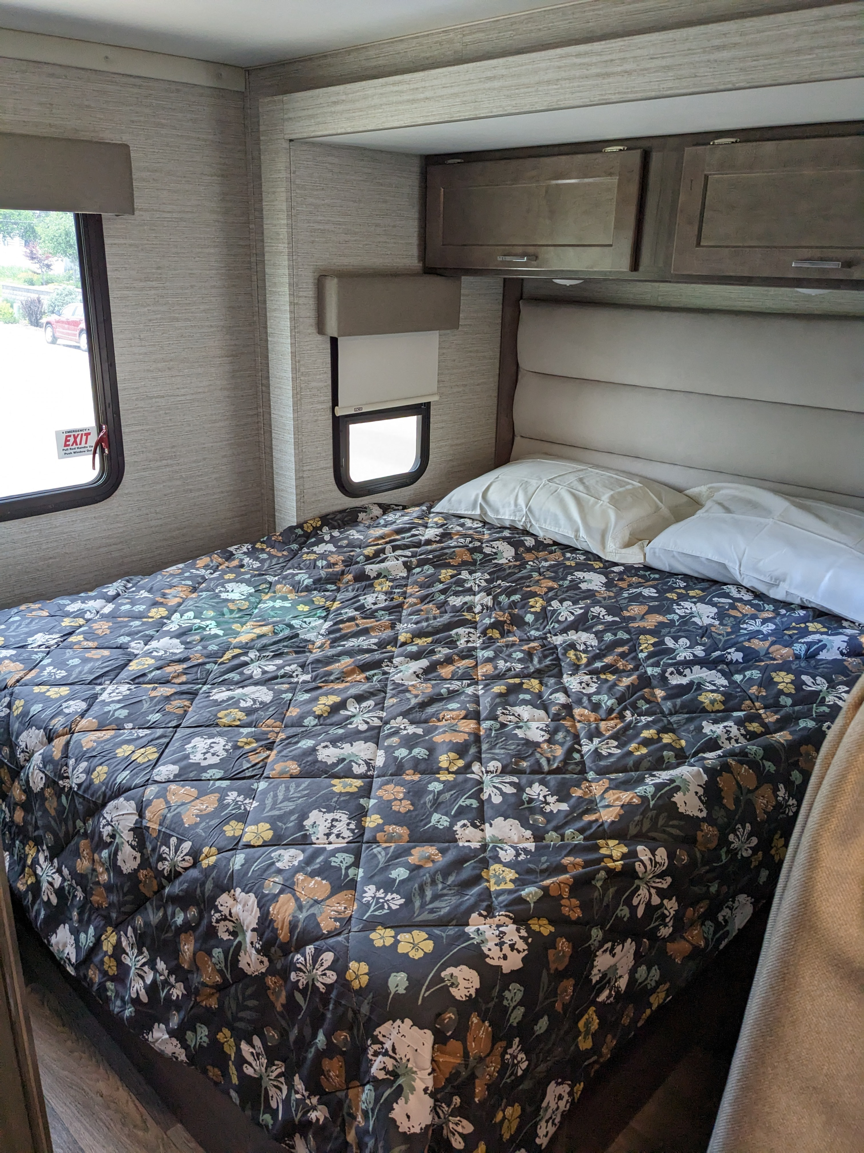Cozy RV rental bedroom features a dark floral comforter, padded headboard, pillows, windows, and overhead cabinets.. Winnebago Minnie Winnie 2022