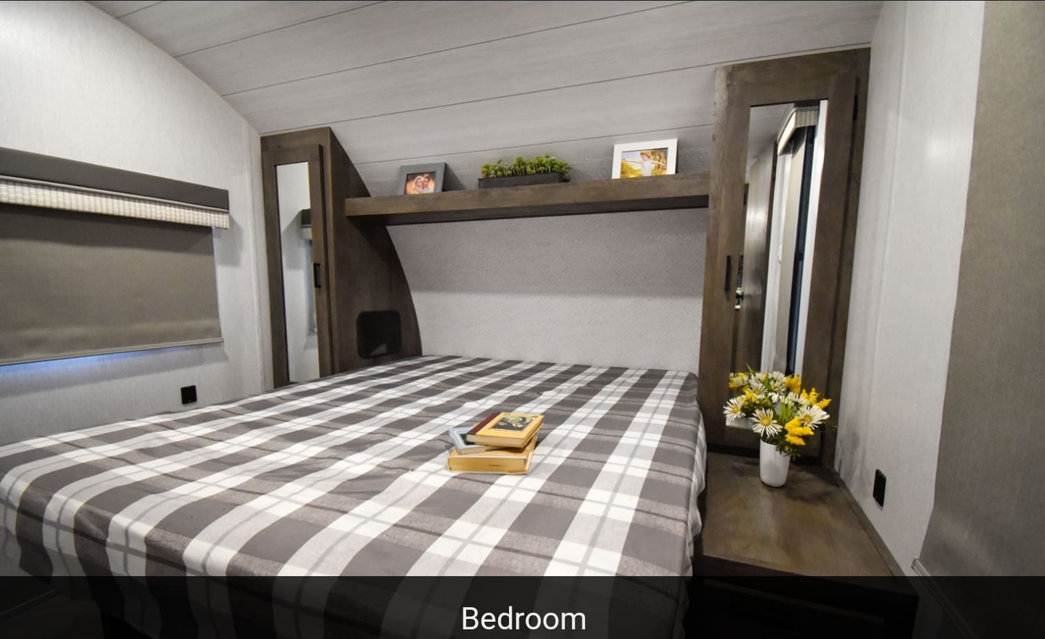 RV rental bedroom with a comfy plaid bed, overhead shelf with decor, and mirrored wardrobe. A cozy travel space.. Forest River Wildwood X-Lite 2023