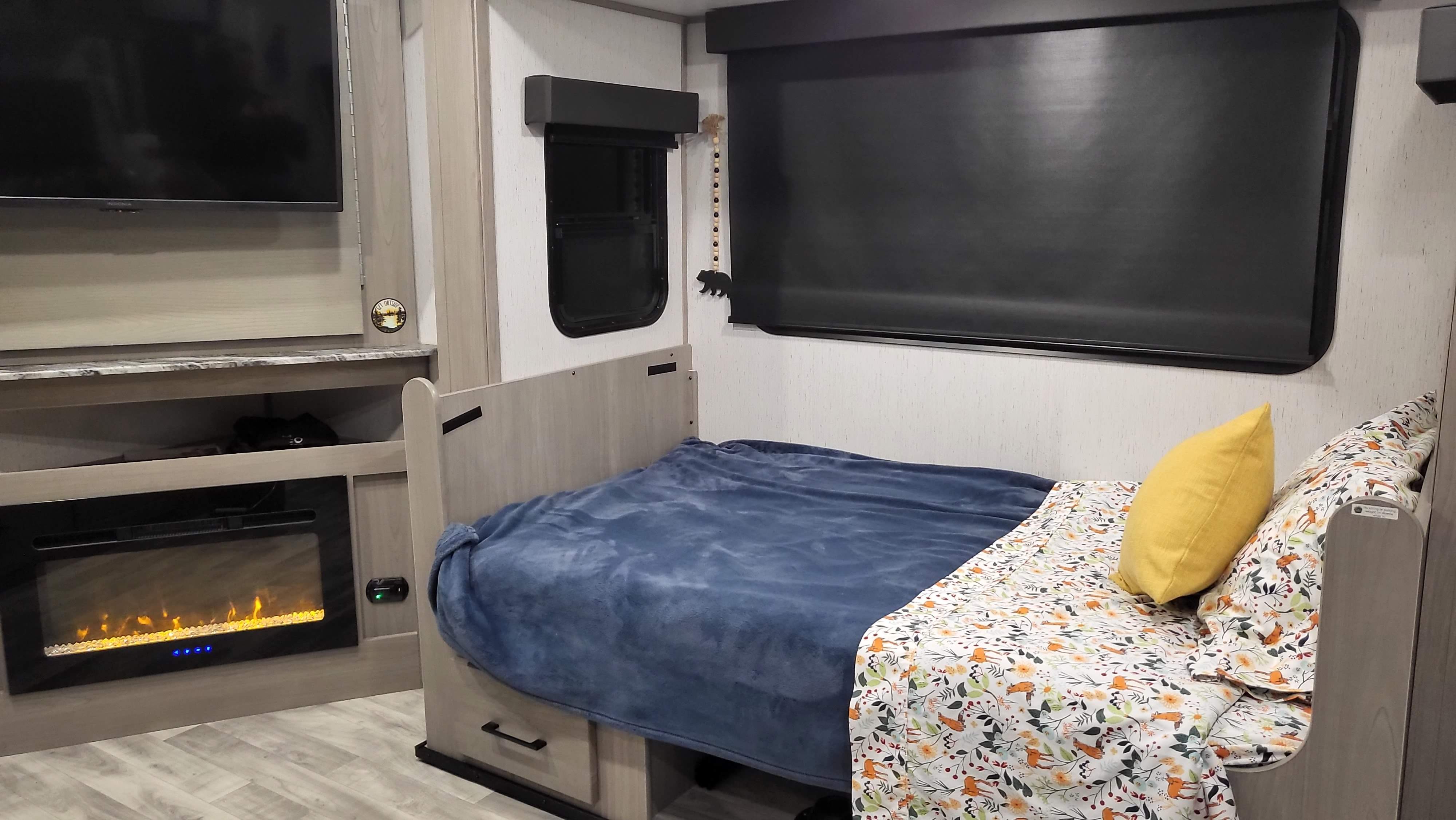Cozy RV rental interior: bed with animal-print sheet, yellow pillow, TV, and electric fireplace. A beaded bear decoration hangs by the window.. Dutchmen Kodiak Ultra Lite 2022