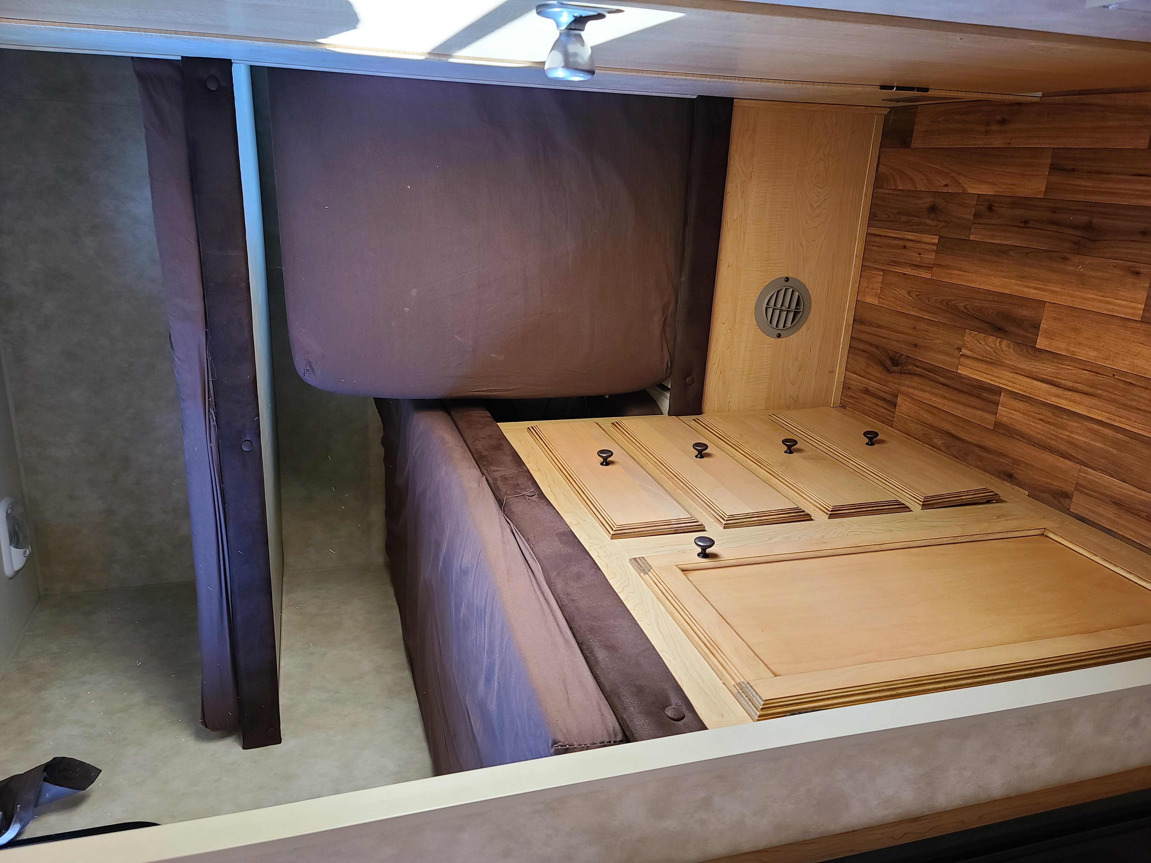 RV rental interior: a bed with wooden lift-up storage panels and knobs, next to a privacy curtain and wood-paneled wall.. Spree Spree Trailer 2010