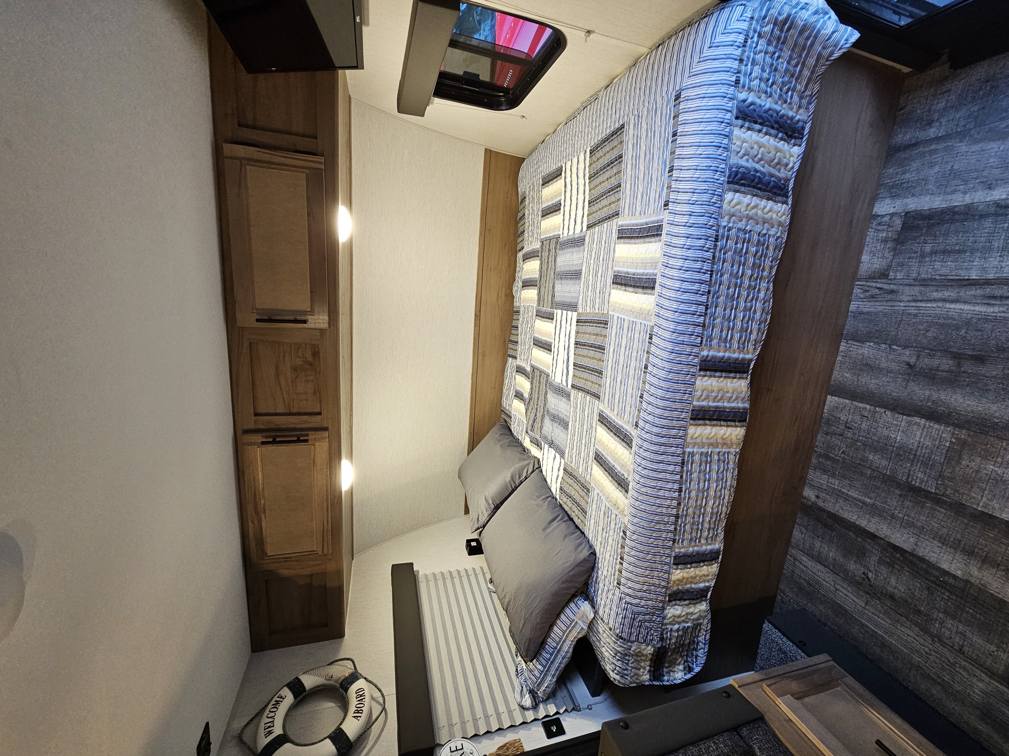 Bright RV rental bedroom: striped quilt on bed, wooden cabinets, overhead window, and a 'Welcome Aboard' lifebuoy.. Forest River Wolf pup 17JW 2023