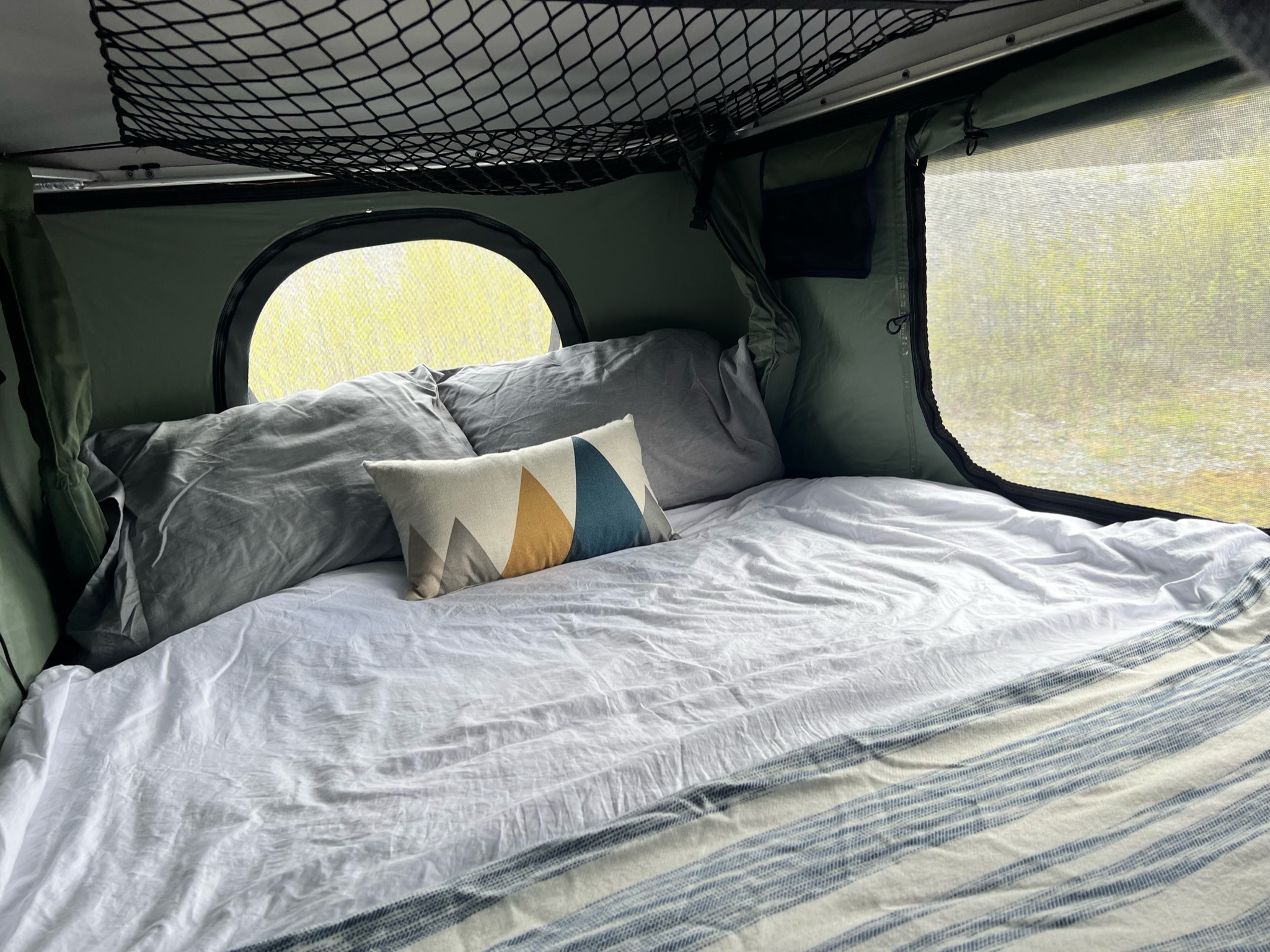 Cozy RV rental interior: a comfy bed with pillows, round window, & mesh screen for serene outdoor views.. Toyota Tacoma 2012