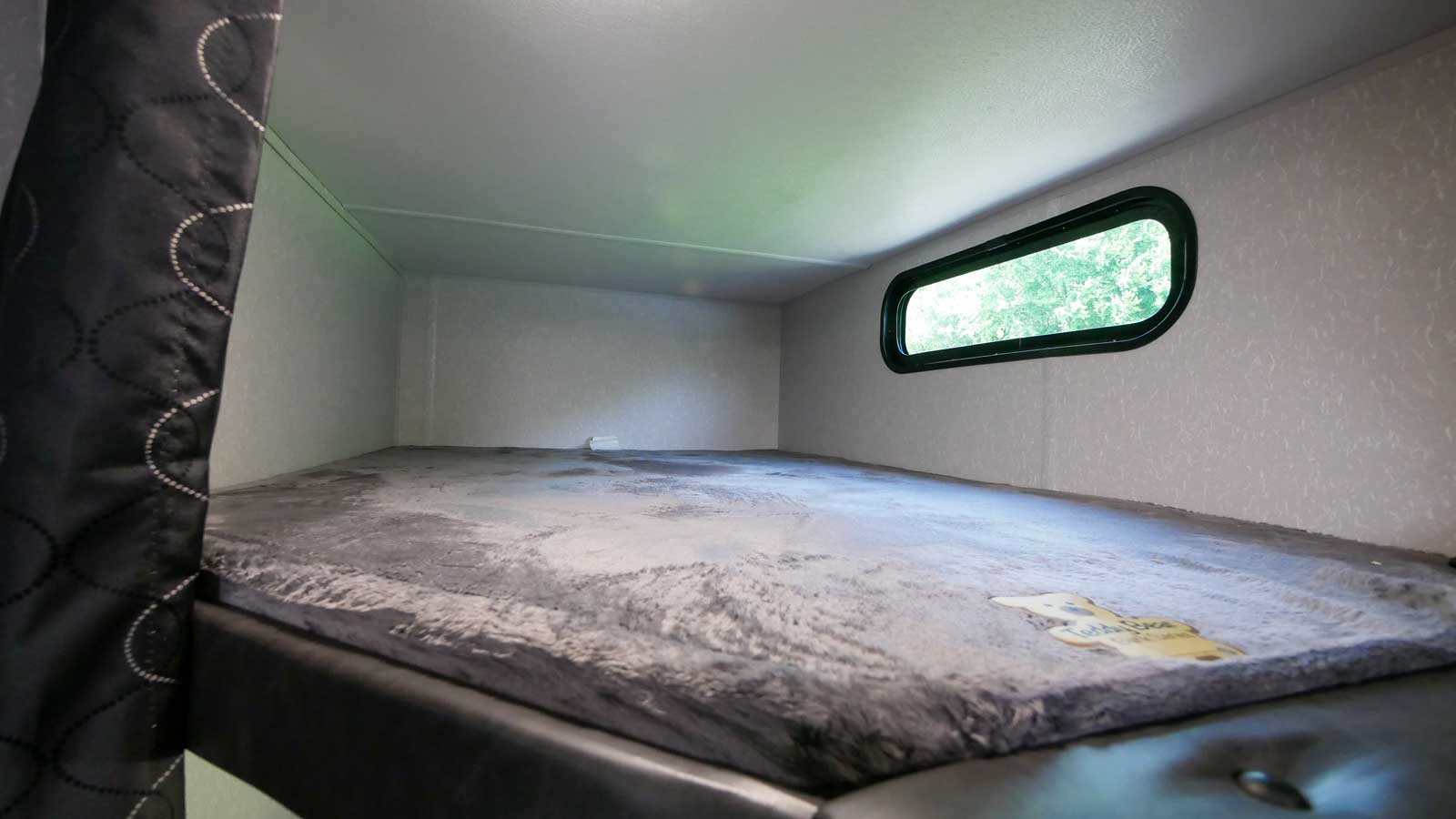 Inside an RV rental, this bunk bed features a grey mattress, a small window with a view of trees, and a dark patterned curtain.. Forest River 271brb 2022