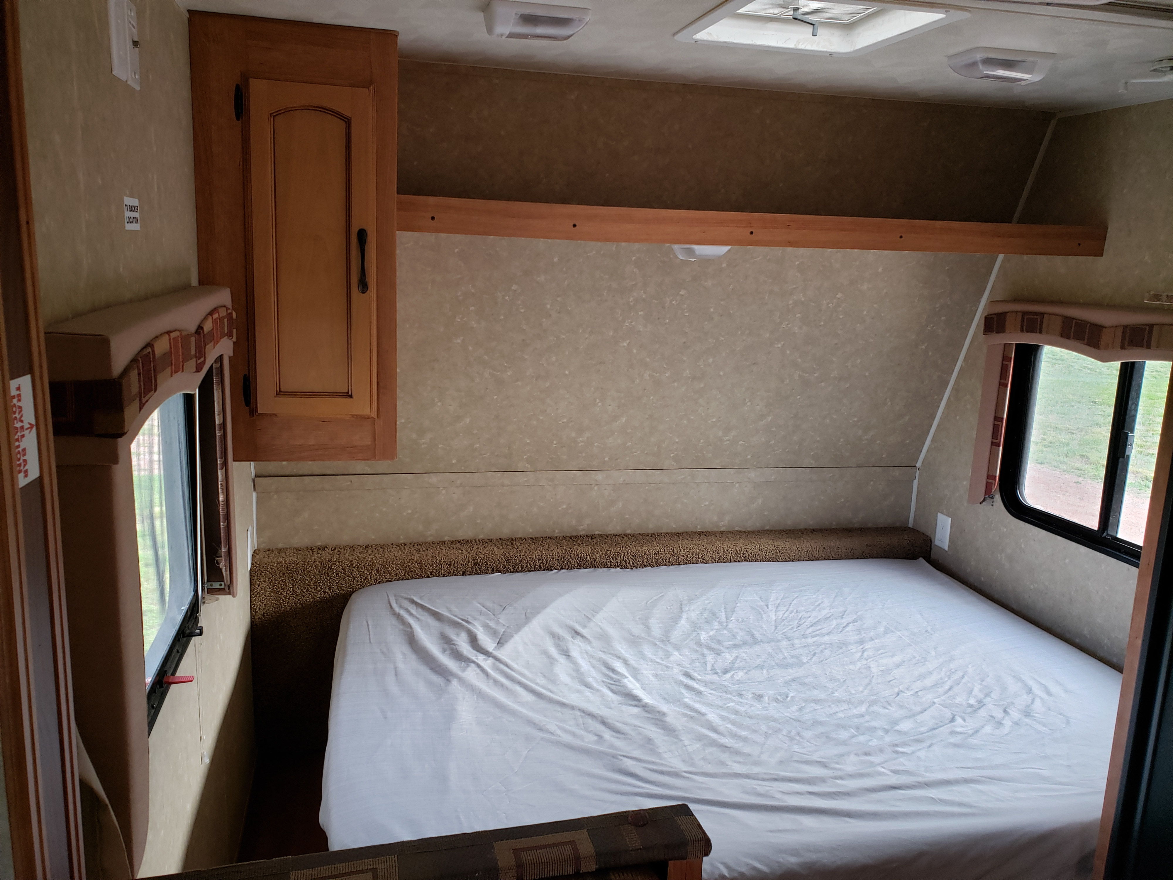 This RV rental features a compact bedroom with a bed, white sheets, side window, and wood cabinetry, ready for a TV.. Gulf Stream Ameri-Lite 2011