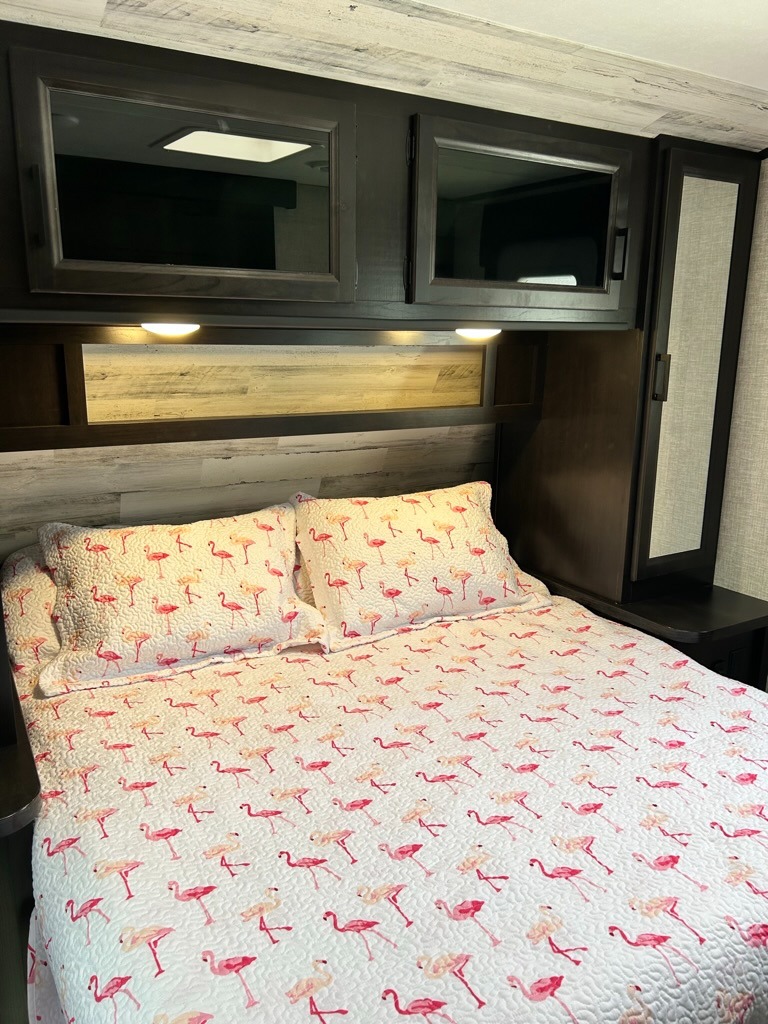 Cozy RV rental bedroom. Features a fun flamingo quilt on the bed, overhead storage cabinets, and soft reading lights.. Jayco Jay Feather 2022