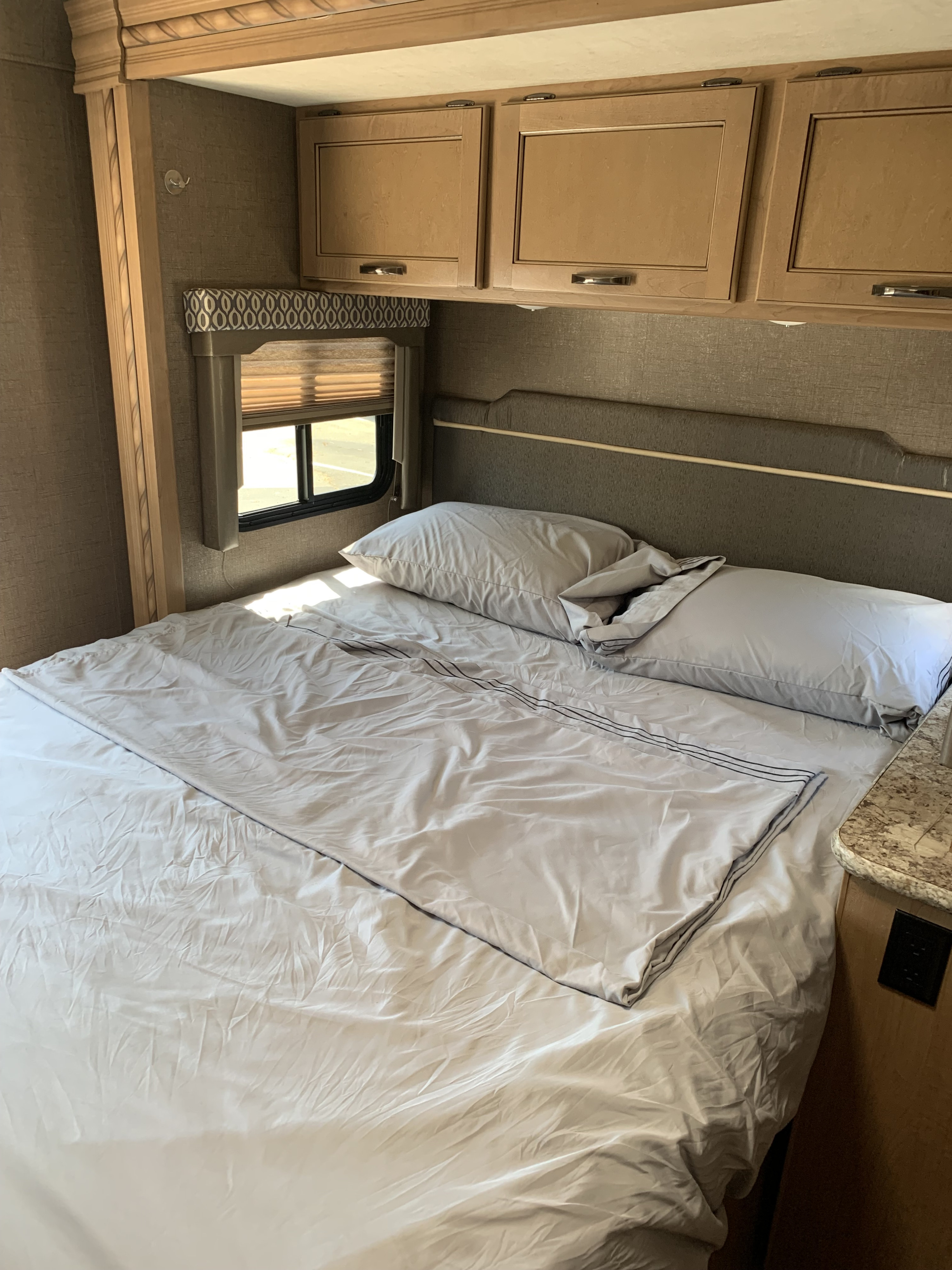 Cozy RV rental bedroom showing a bed with light grey linens, overhead cabinets, and a window for comfortable travel.. Thor Freedom Elite 2019