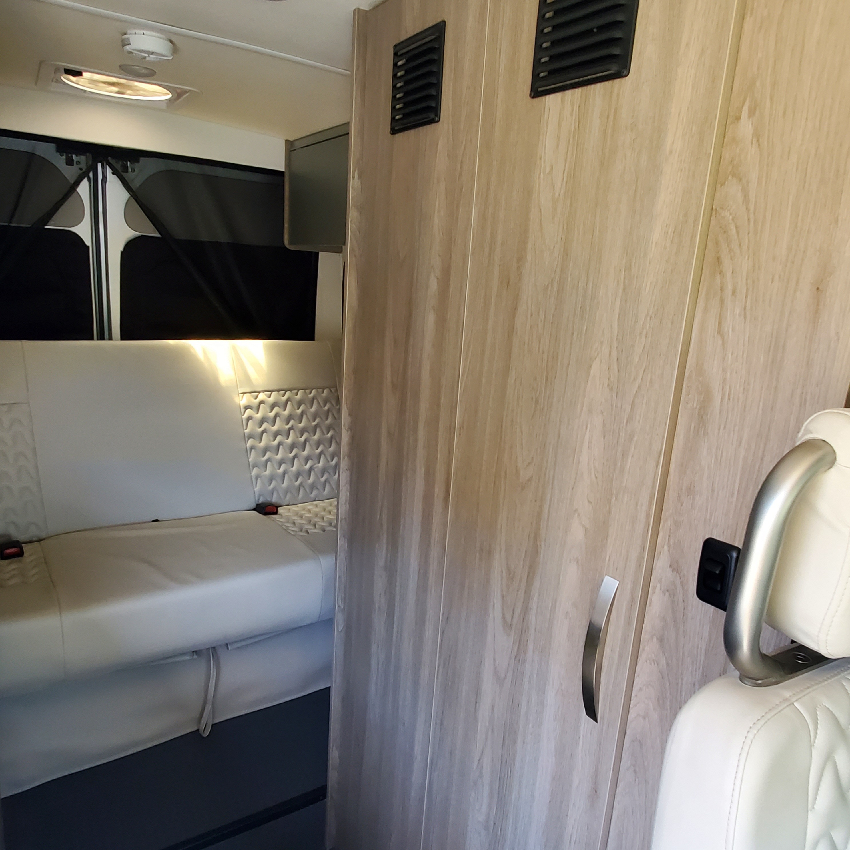 Modern RV rental interior featuring light wood cabinets, white upholstered seating with seatbelts, and ceiling light.. Winnebago Solis 2022