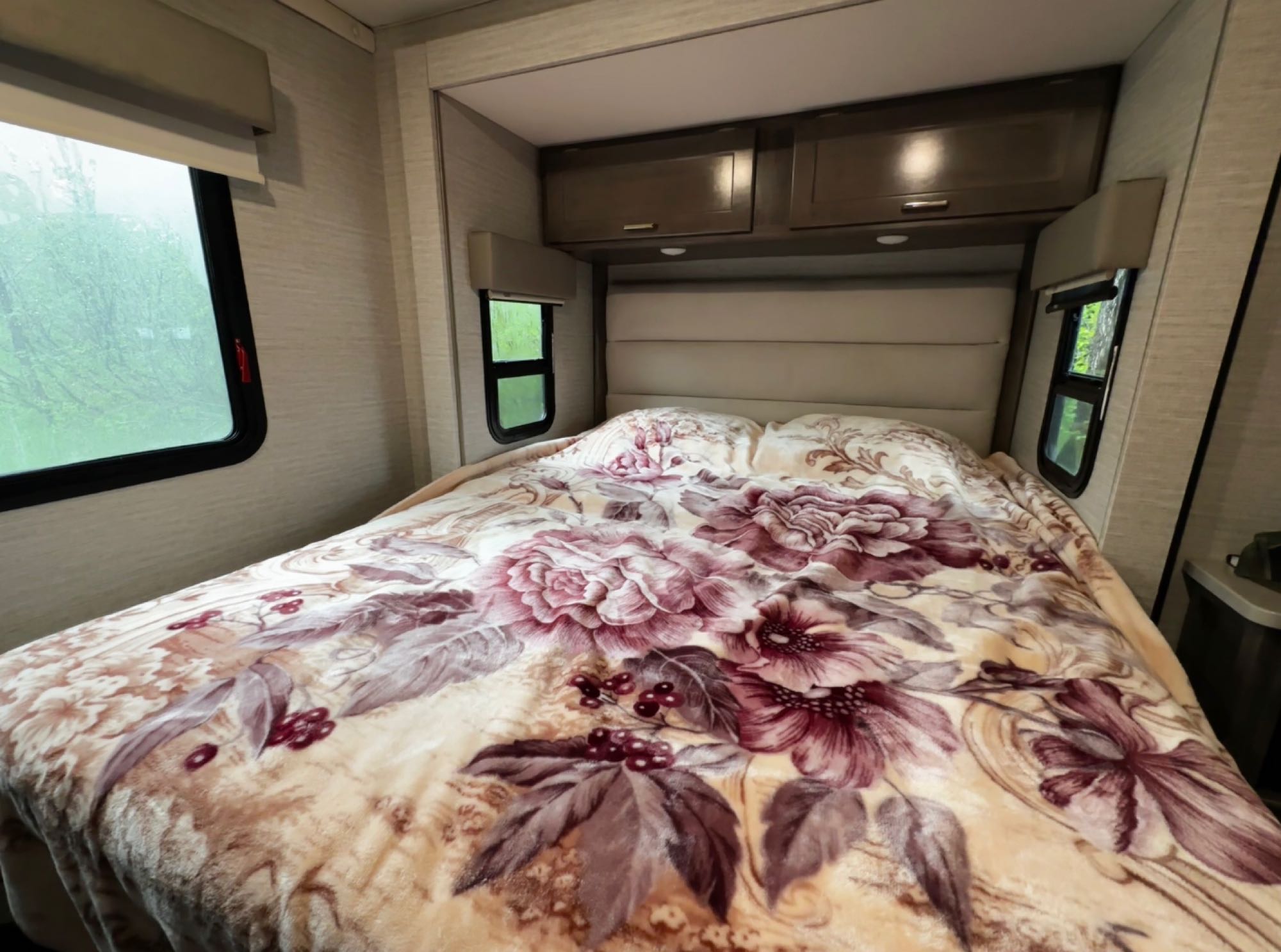 Cozy RV rental bedroom with a floral blanket, padded headboard, and windows showing outdoor greenery.. Winnebago Minnie Winnie 2020
