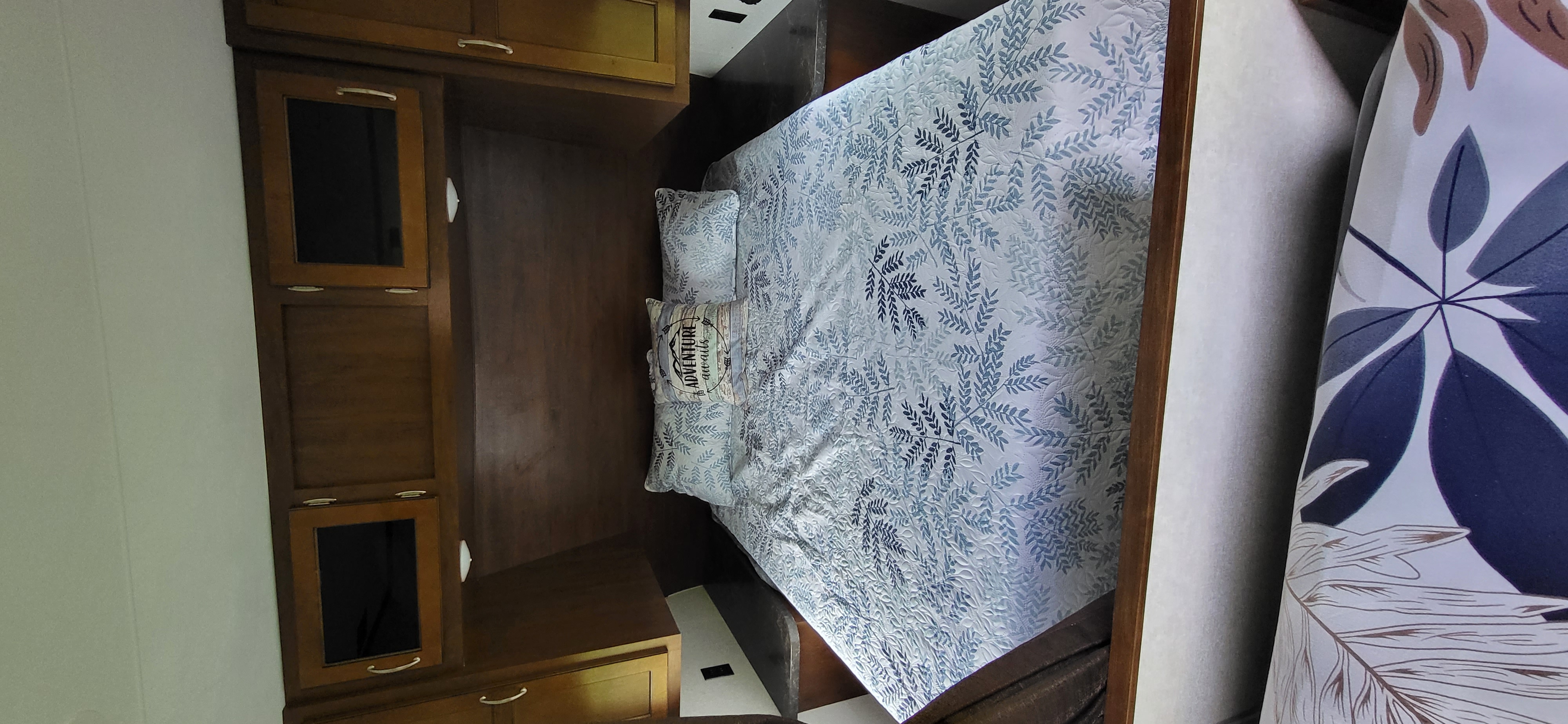 Cozy RV rental bedroom features a bed with blue leaf-patterned bedding, an 'ADVENTURE awaits' pillow, and wood cabinets.. Jayco Jay Flight 2017