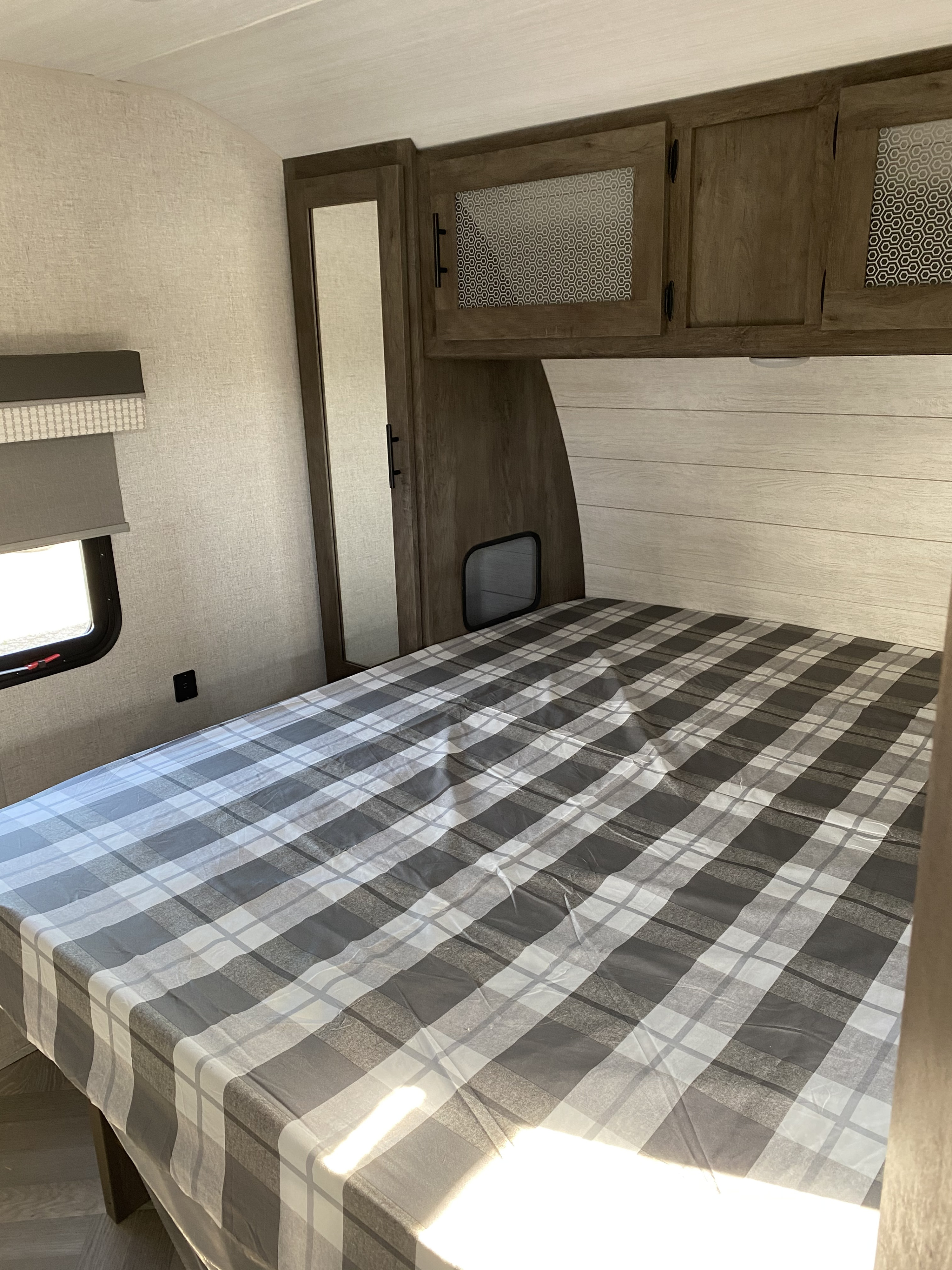Cozy RV rental bedroom featuring a plaid bed, overhead wooden cabinets, and a side window with a shade.. Forest River Wildwood 2021