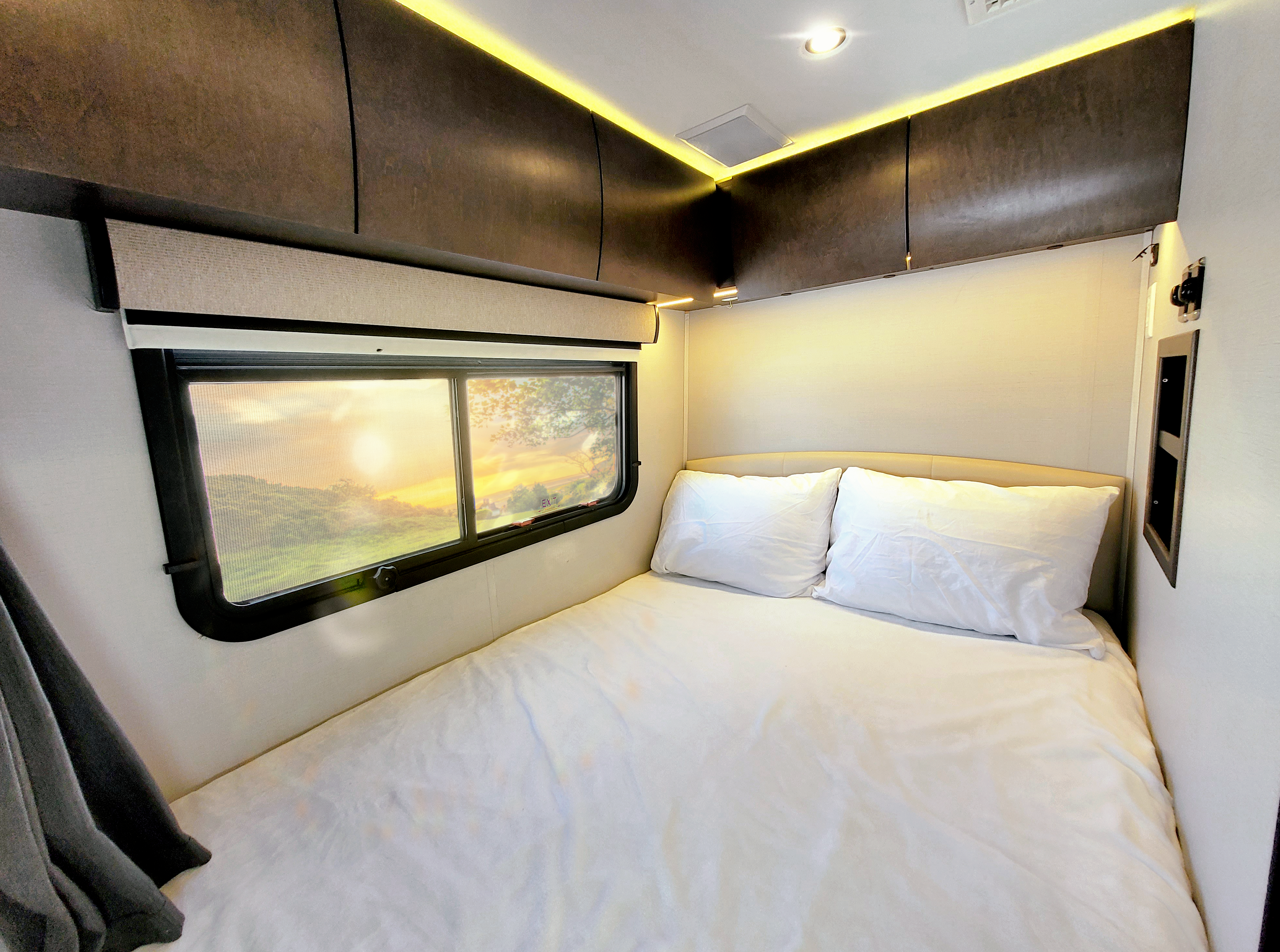 RV rental: Bright bedroom with a large bed, two white pillows, overhead cabinets, and a window displaying a vibrant sunrise or sunset.. Leisure Travel Unity 2018