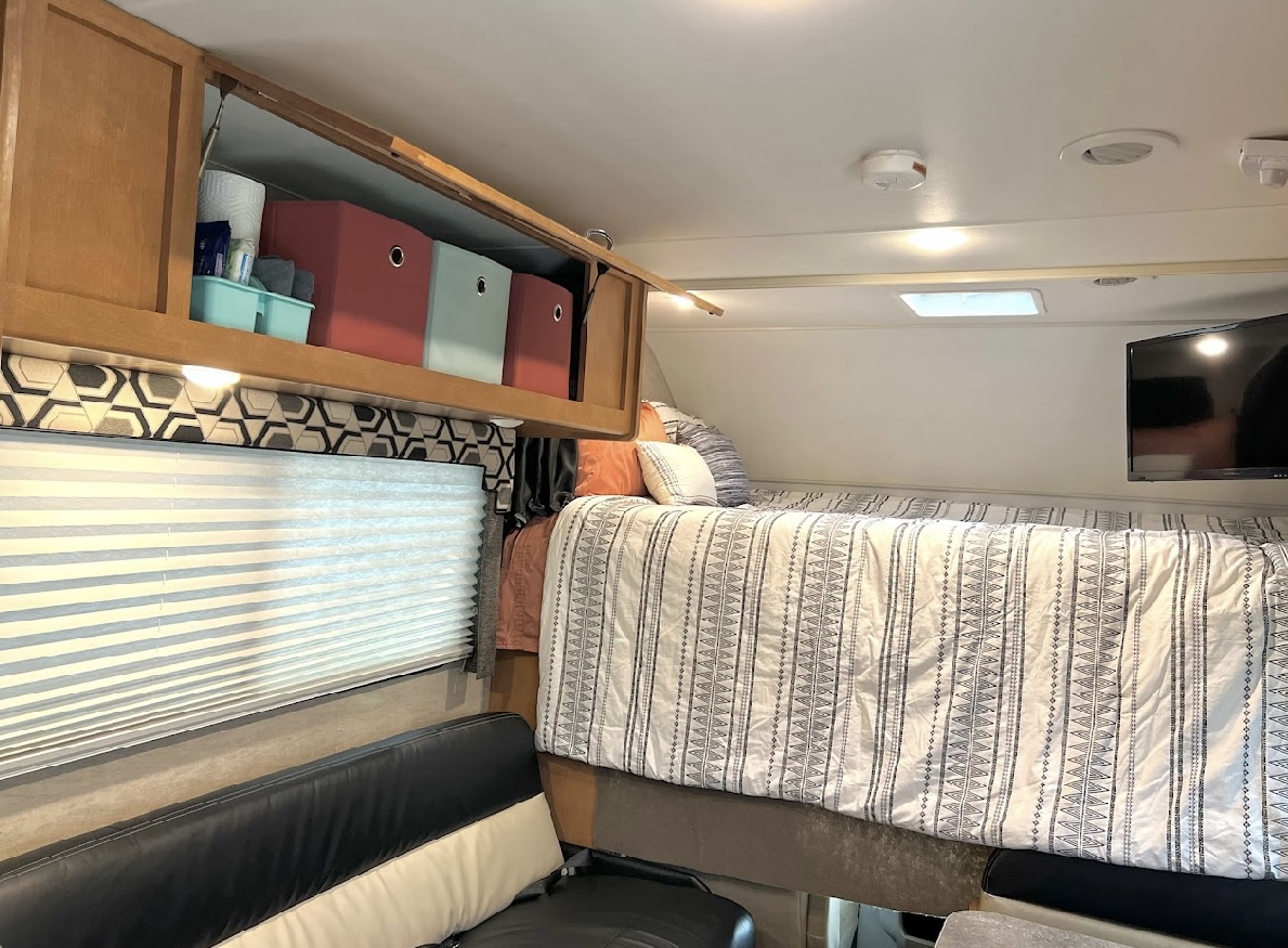 RV rental interior: View of the cab-over bed with patterned bedding, open overhead storage, a TV, window, and seating below.. Winnebago Minnie Winnie 2017