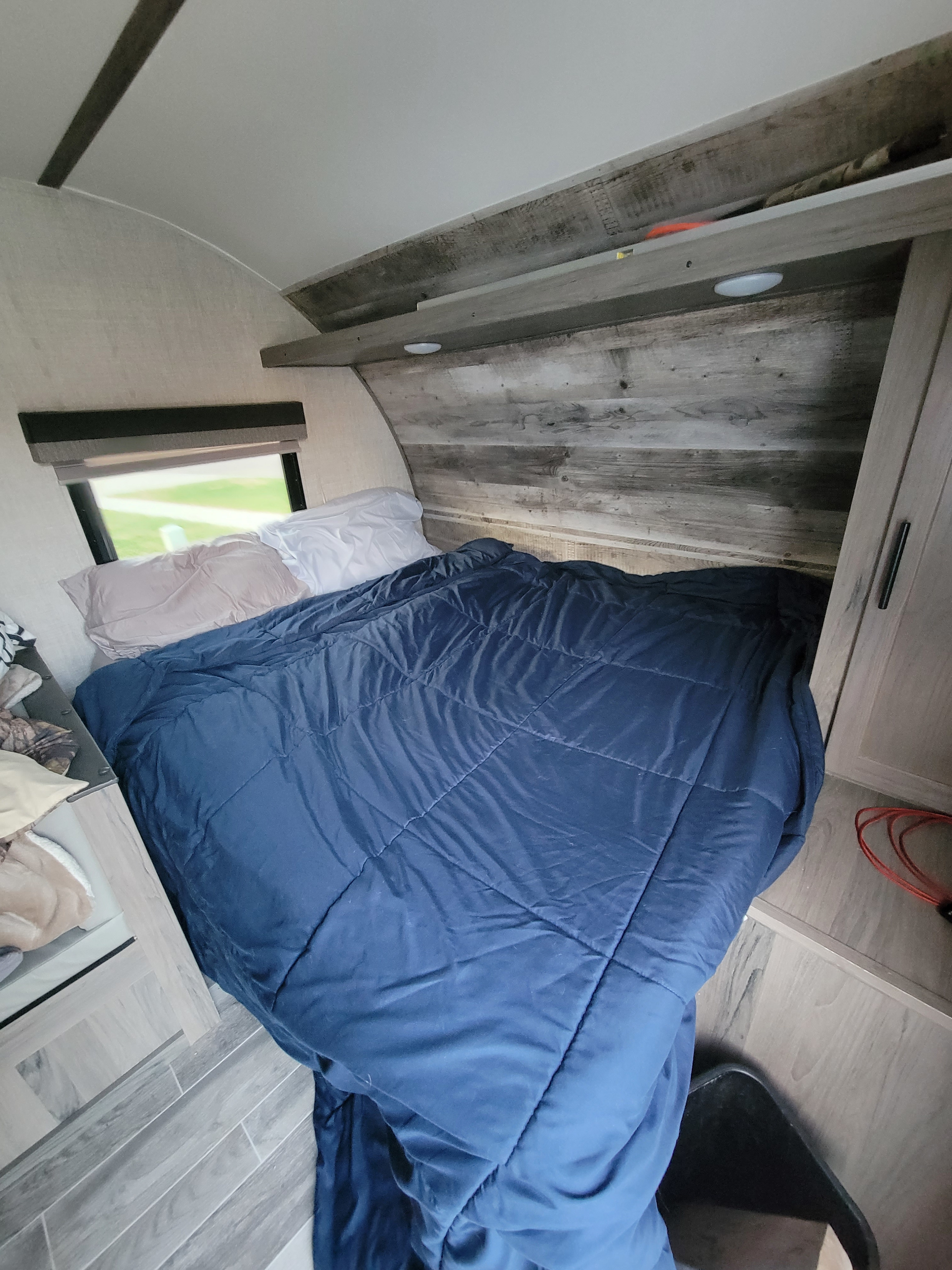 Inside an RV rental: a cozy bedroom with a large bed, blue comforter, pillows, window view, and wooden storage.. Gulf Stream Ameri-Lite Super Lite 2021