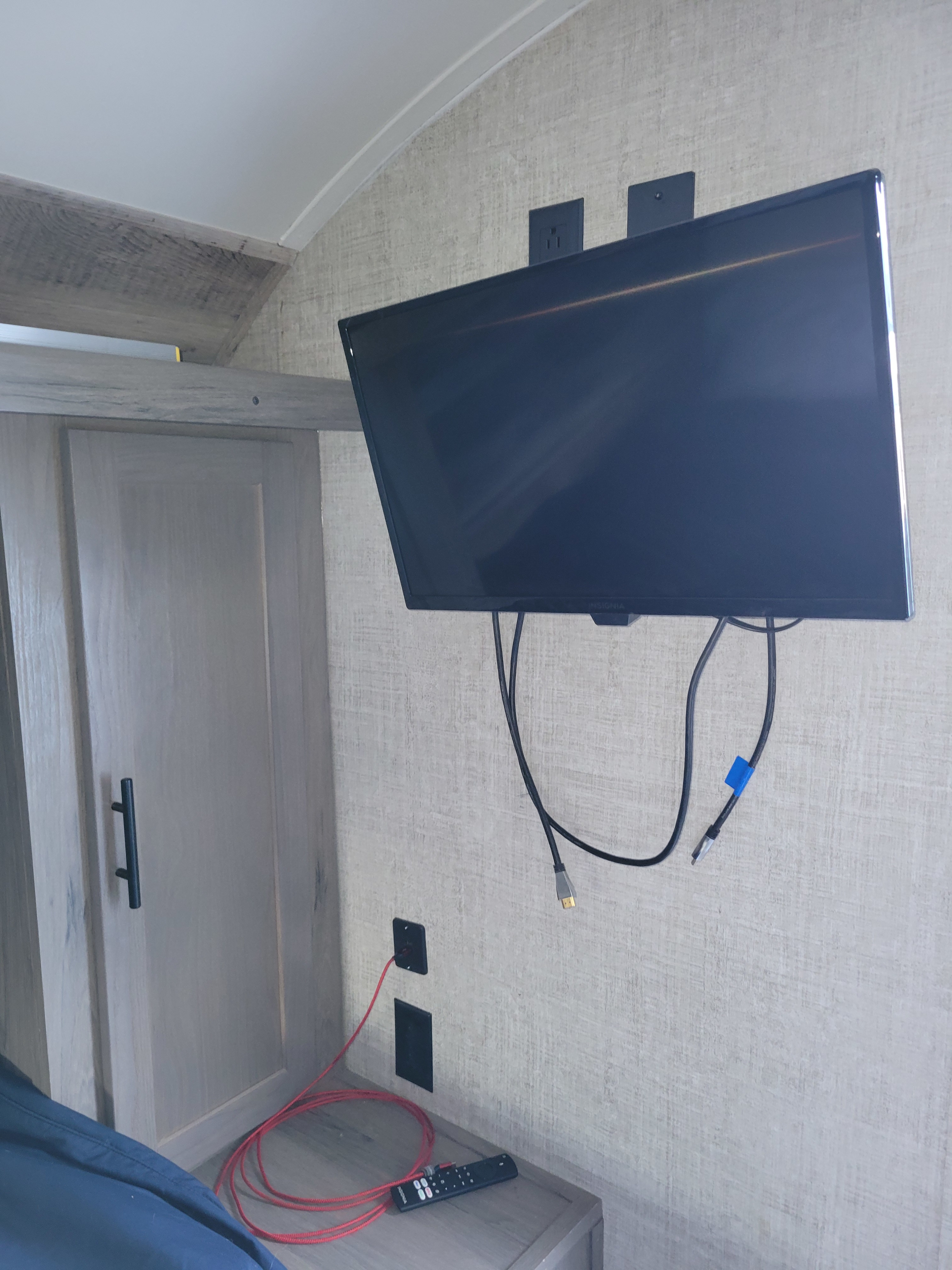 RV rental bedroom: Wall-mounted TV with dangling cables, outlets, and remote on nightstand next to blue bedding.. Gulf Stream Ameri-Lite Super Lite 2021