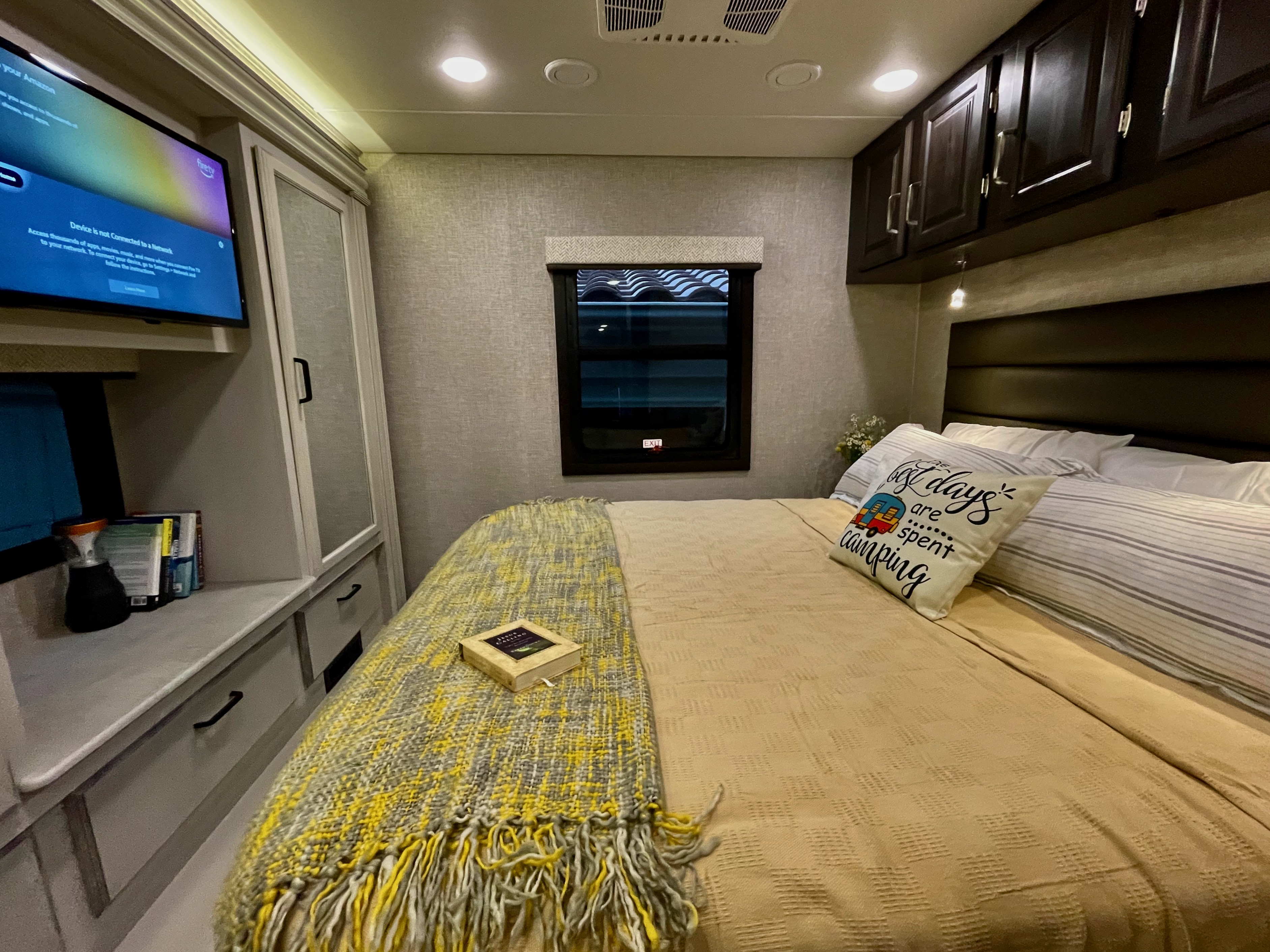 Inside a cozy **RV rental** bedroom showing a comfortable bed with a "camping" pillow, TV, and storage cabinets.. Entegra Coach Vision XL 2023