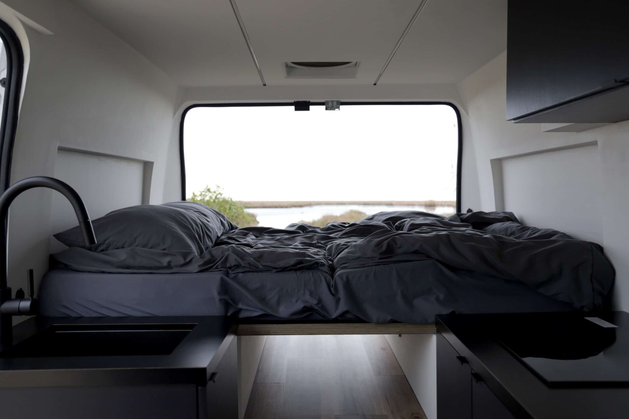 RV rental interior: A cozy dark bed at the rear overlooks a serene waterscape, with sleek black kitchen counters below.. RAM Promaster 2018