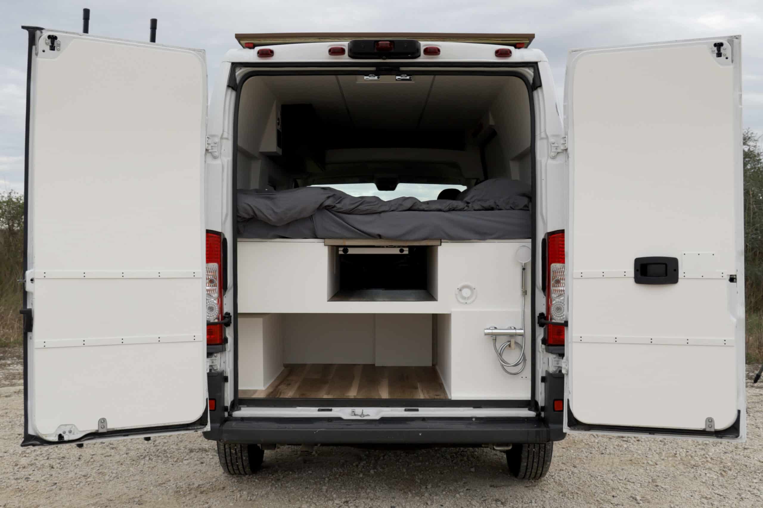 Explore this white camper van RV rental, showcasing its open rear doors revealing a cozy bed, integrated storage, and an outdoor shower.. RAM Promaster 2018