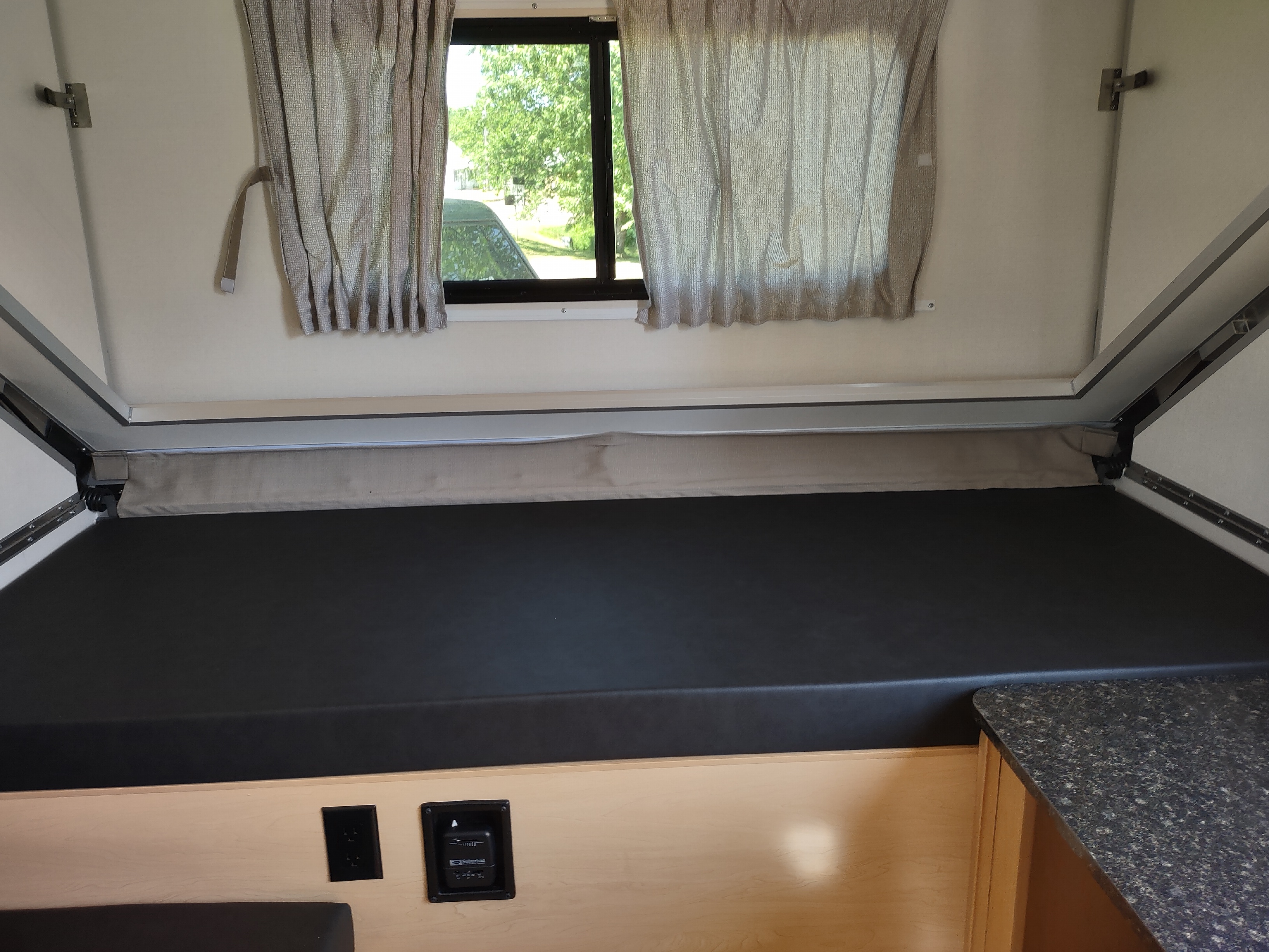 RV rental: Interior of a pop-up camper showing a bed, window with curtains, control panel, & small counter. Ideal for travel.. A Liner A Liner Popup Trailer 2022