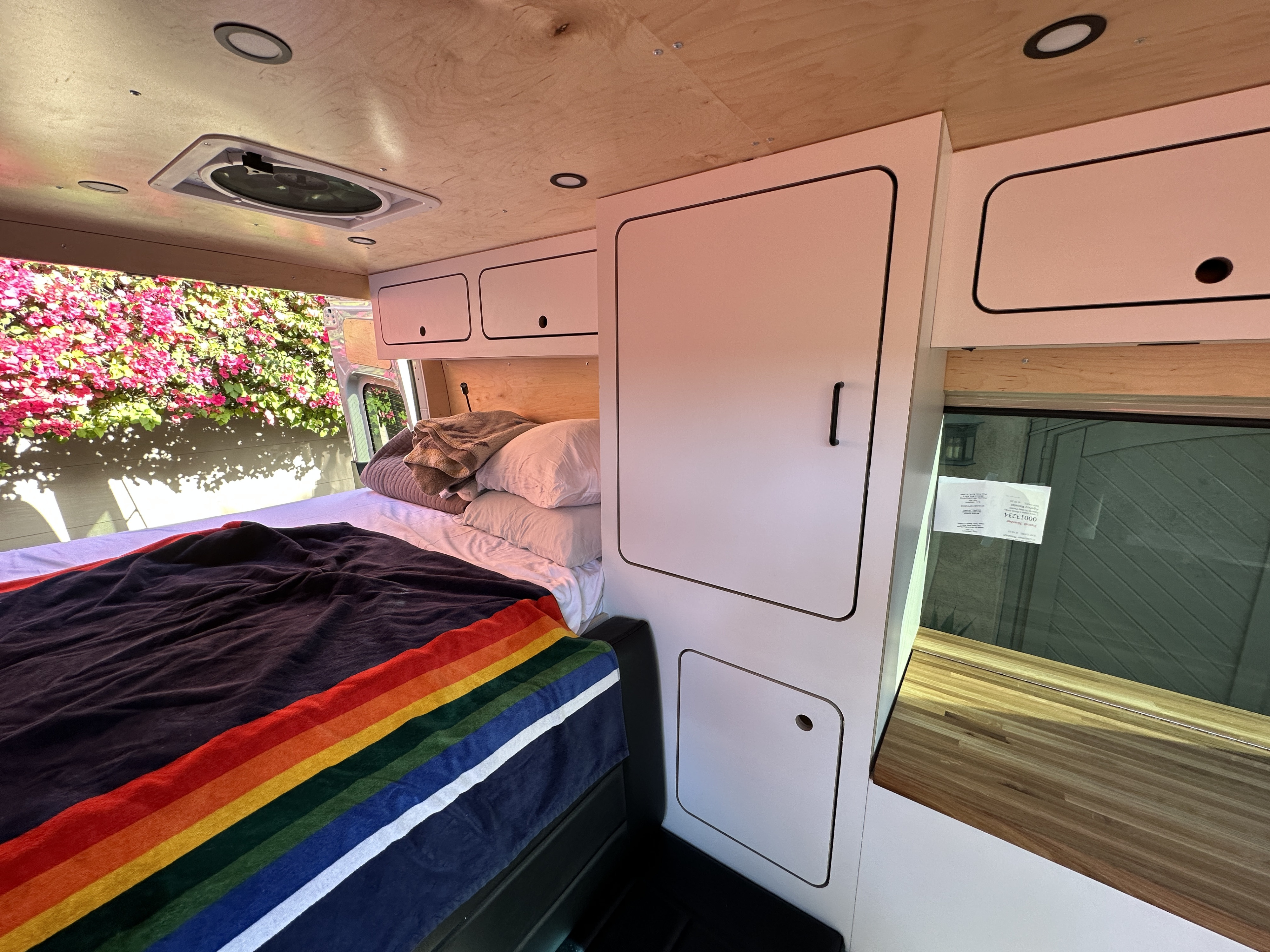Cozy camper van interior for your next RV rental, featuring a bed, storage, and natural light from a window overlooking flowers.. VanCraft VanCraft Class B 2023
