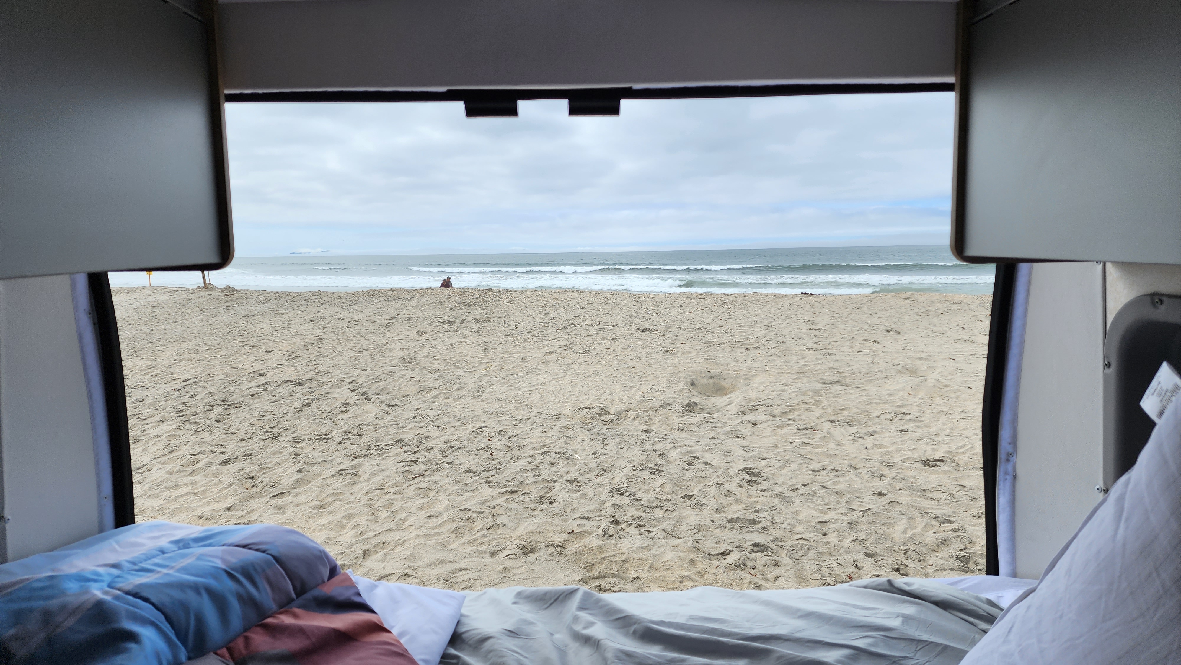 Relaxing RV rental view of a sandy beach & ocean, seen from inside with cozy bedding. A distant person by the waves.. Winnebago Solis 2023