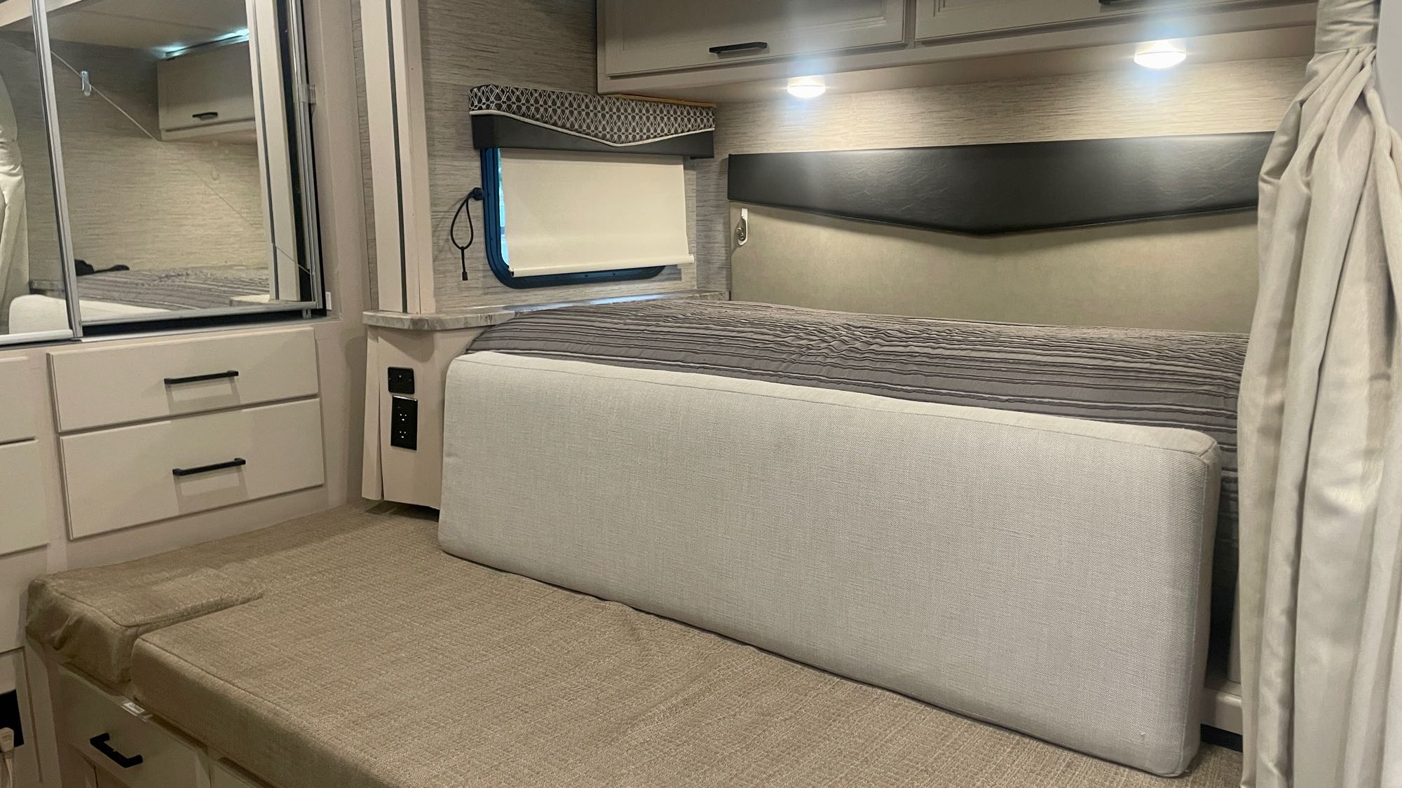 Inside a bright RV rental: view of a bedroom with bed, built-in mirrored storage, seating, and window with blind.. Thor Compass 2021