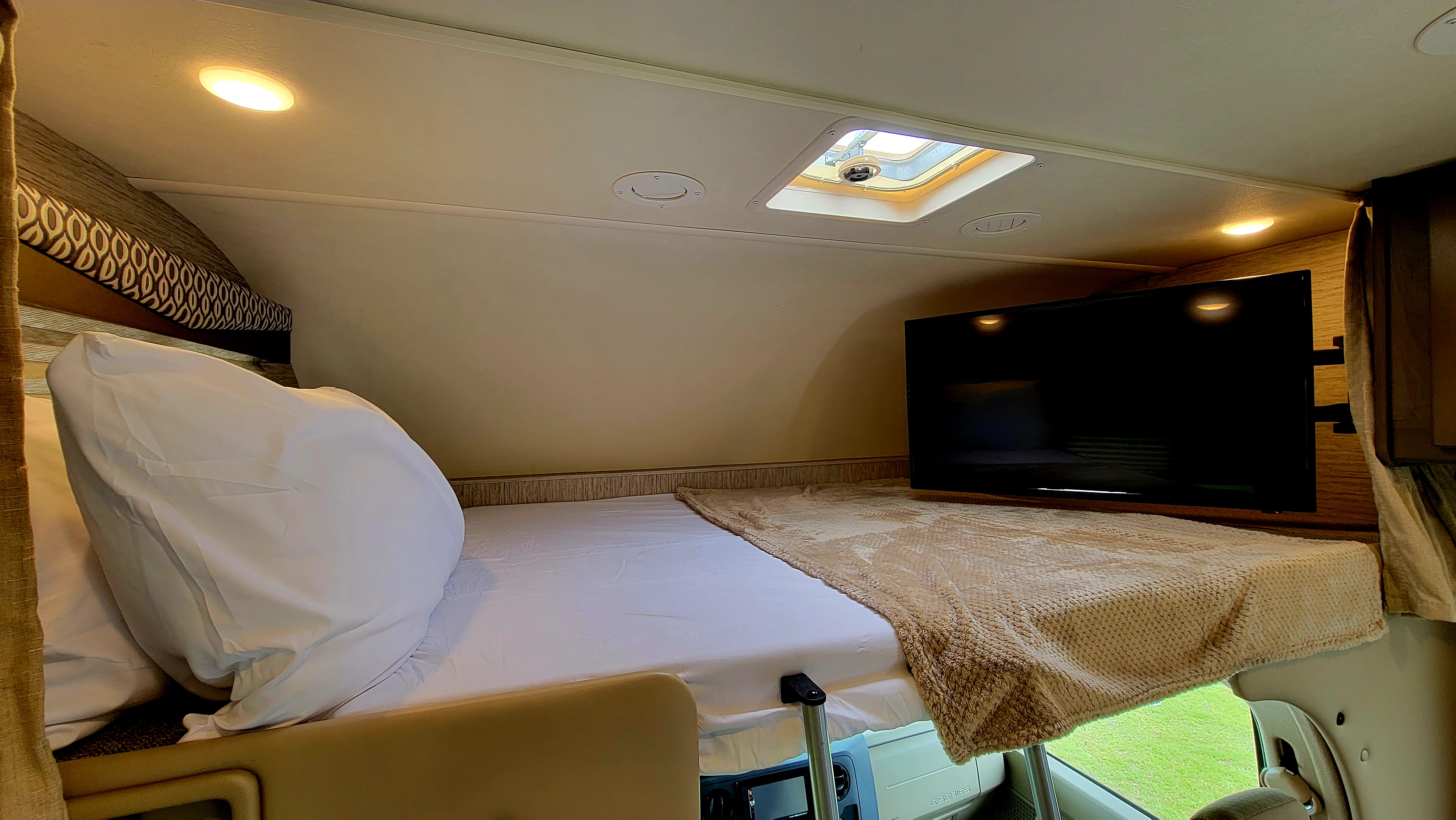Cozy over-cab bunk bed in an RV rental, featuring a comfortable bed, TV, and bright lighting for restful trips.. Thor Freedom Elite 2021