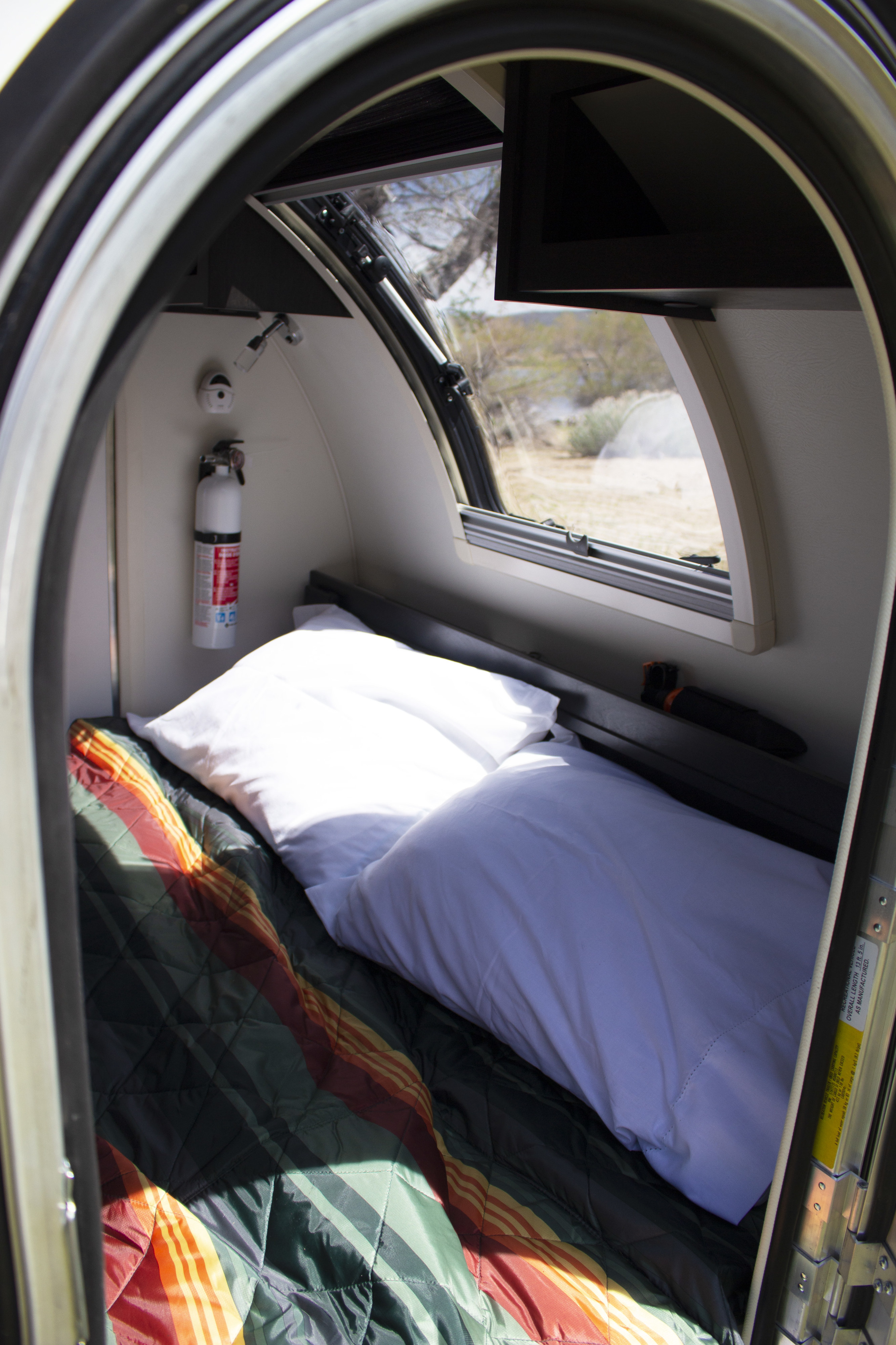Inside a compact RV rental: a cozy bed, fire extinguisher, and window with scenic desert views.. nuCamp T@G 2022