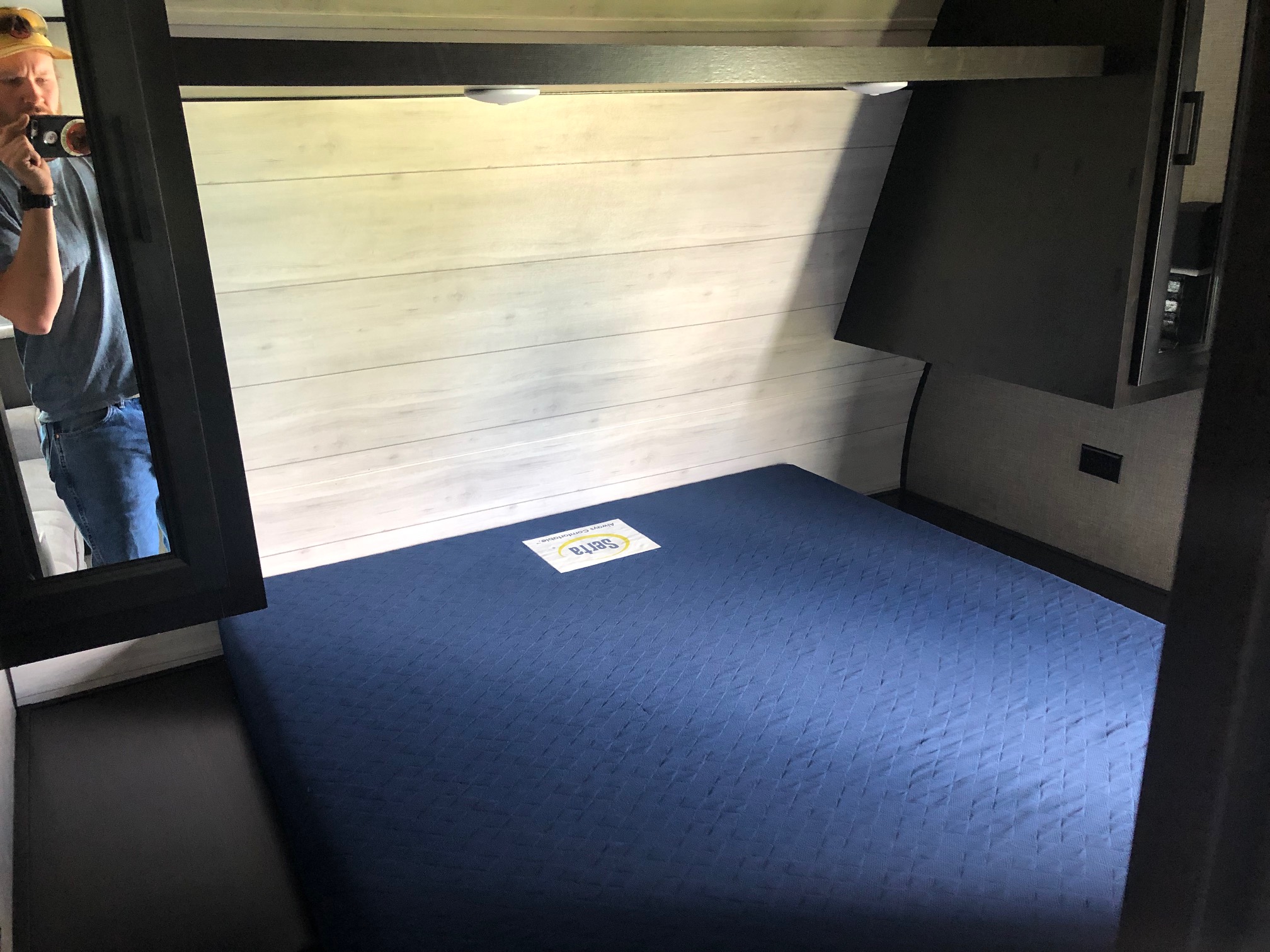 Inside an RV rental, a compact bedroom features a blue Serta mattress, light paneled walls, overhead lights, and mirror reflection.. Jayco Jay Flight 2022