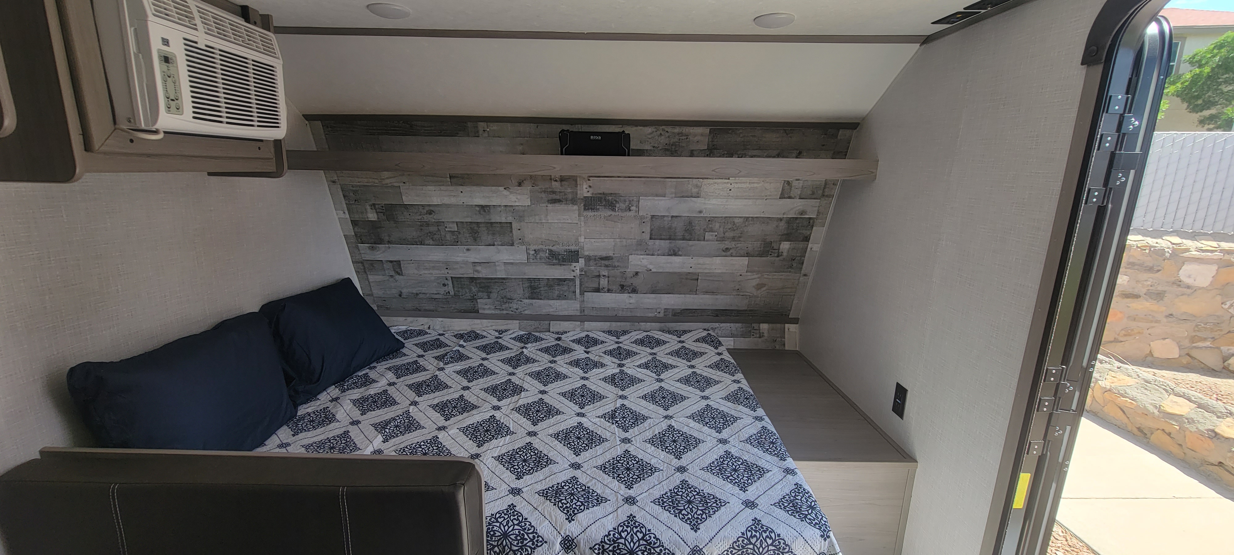 Inside an RV rental, a compact bedroom features a patterned bed, rustic plank wall, A/C, and a BOSS speaker.. Dutchmen Coleman Lantern LT 2023