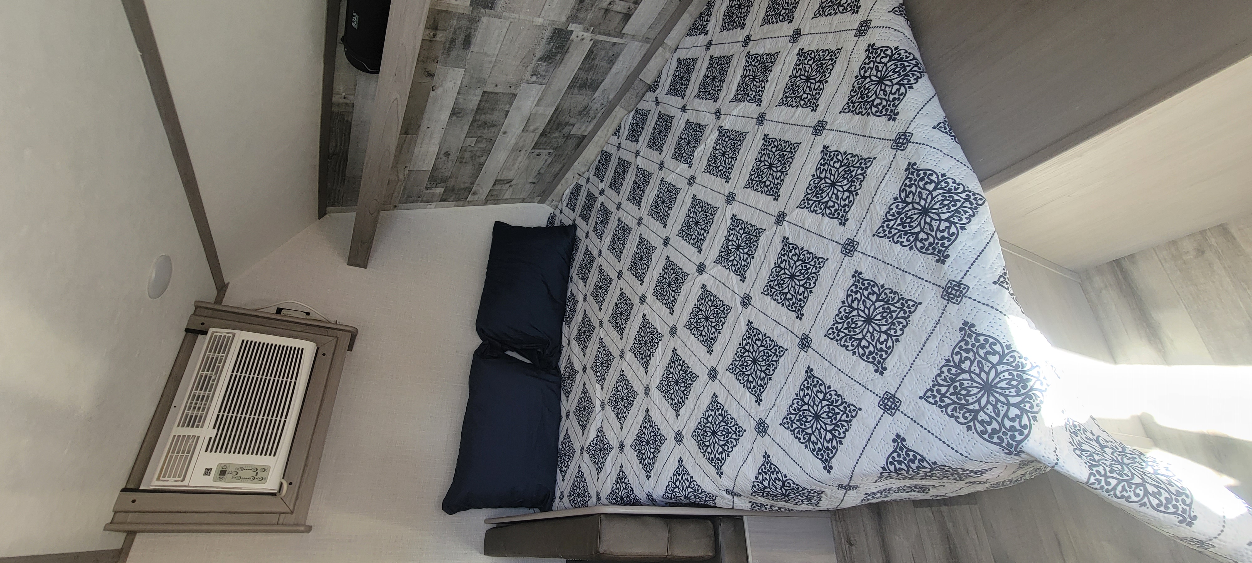 Inside a comfortable RV rental: bed with blue/white quilt, dark pillows, wood paneling, and wall AC unit. Sunny.. Dutchmen Coleman Lantern LT 2023