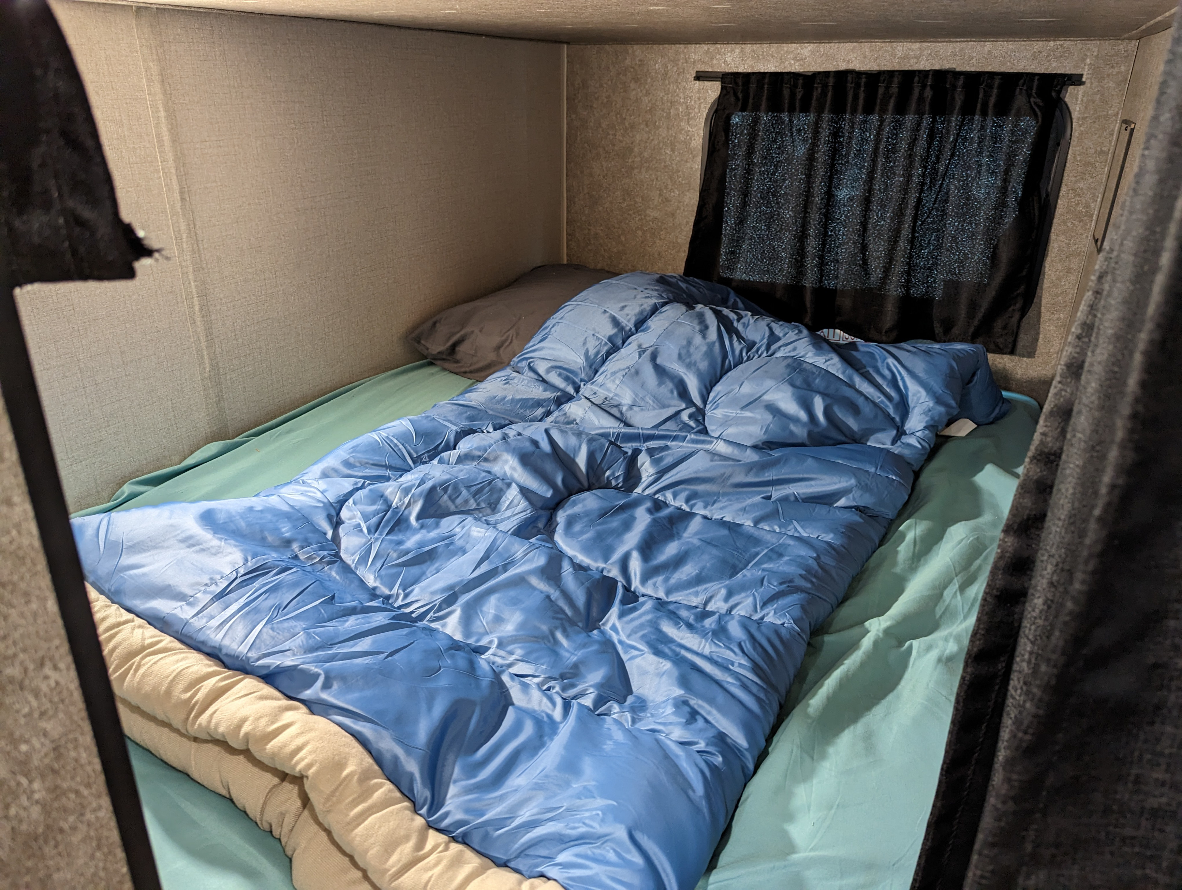 Cozy RV rental bunk bed area with a blue sleeping bag, dark pillow, light green sheets, and window curtain.. Starcraft Autumn Ridge 2021