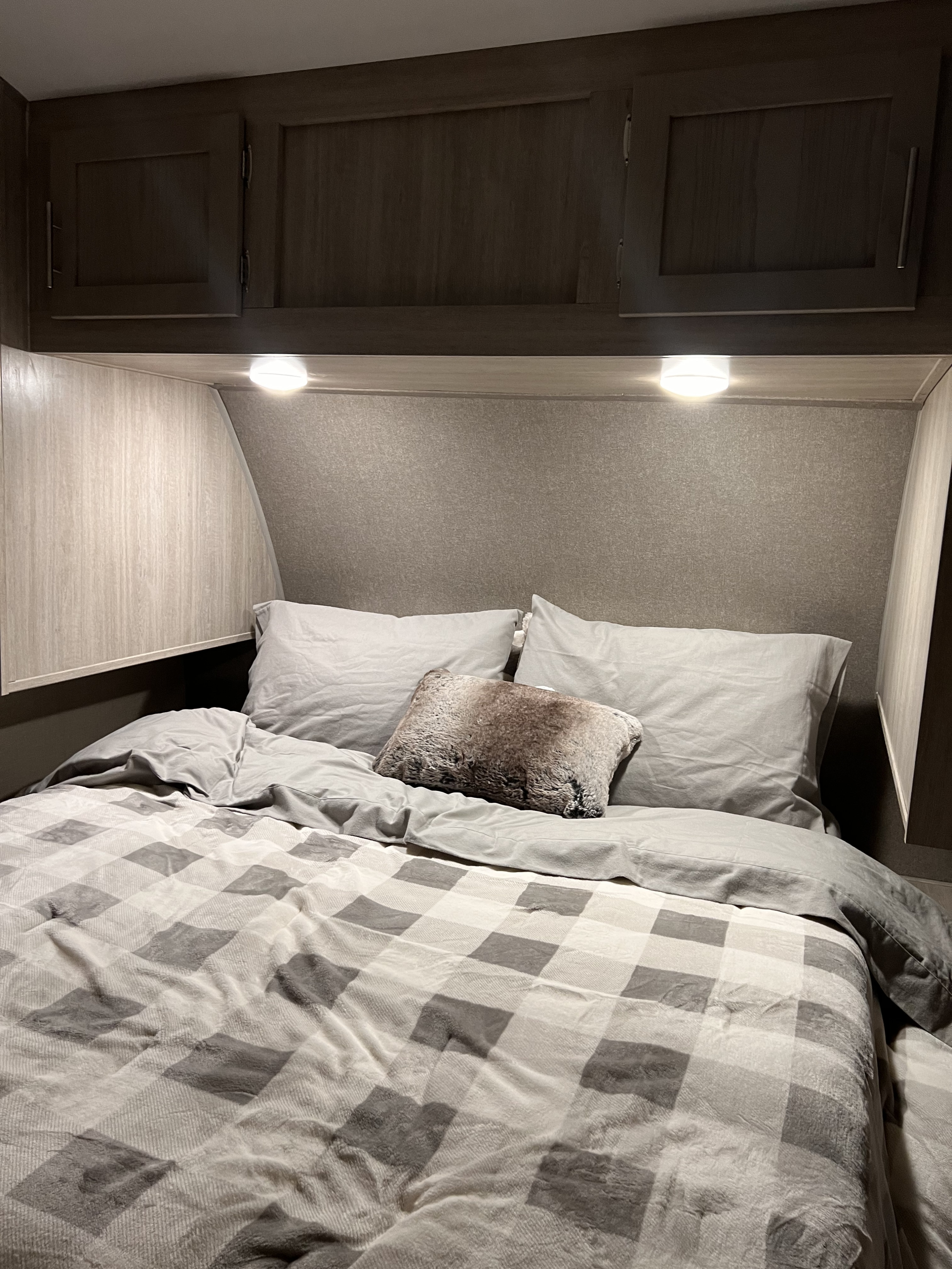 View of a modern RV rental bedroom with dark wood cabinets, under-cabinet lighting, and a cozy gray plaid bed.. Coachmen Catalina Legacy Edition 2022