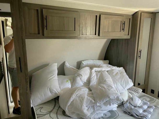 RV rental bedroom: unmade bed with white linens, overhead cabinets, and a person standing in the doorway.. Forest River Shasta 2021