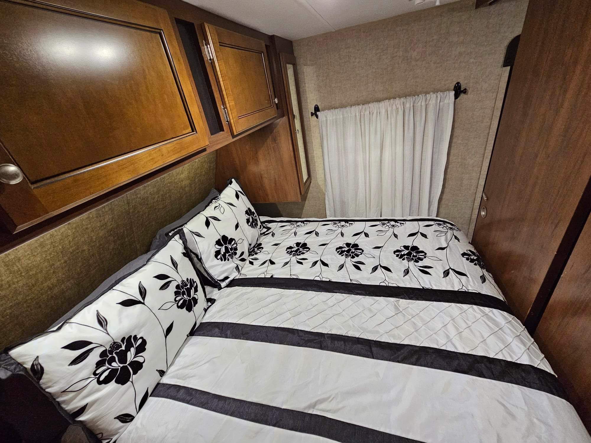 Cozy RV rental bedroom featuring a bed with black & white floral bedding, overhead cabinets, and wall curtains.. Heartland Prowler 2015