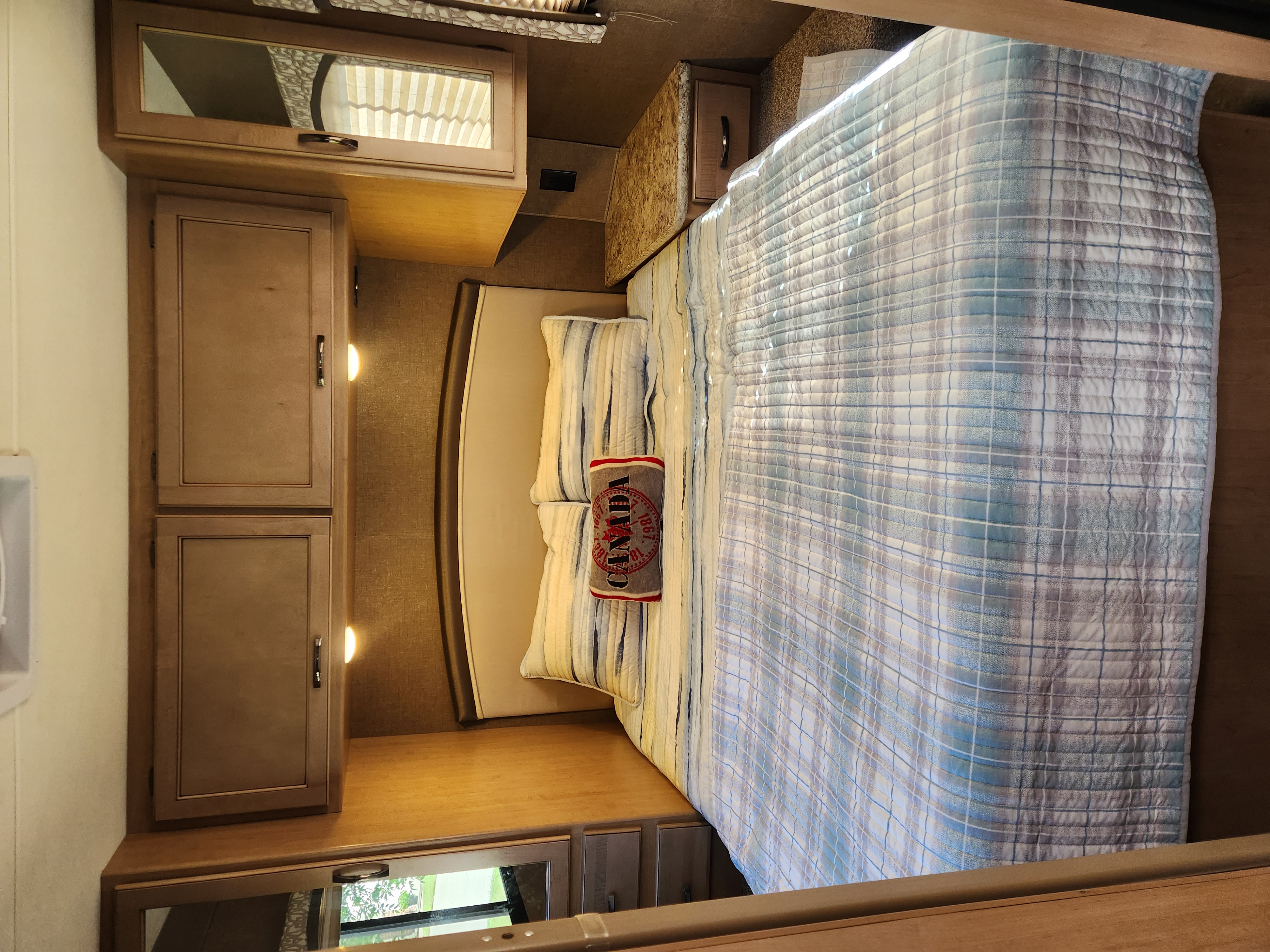 Cozy RV rental bedroom featuring a bed with plaid comforter, pillows, and wooden storage cabinets.. Thor Chateau 2019