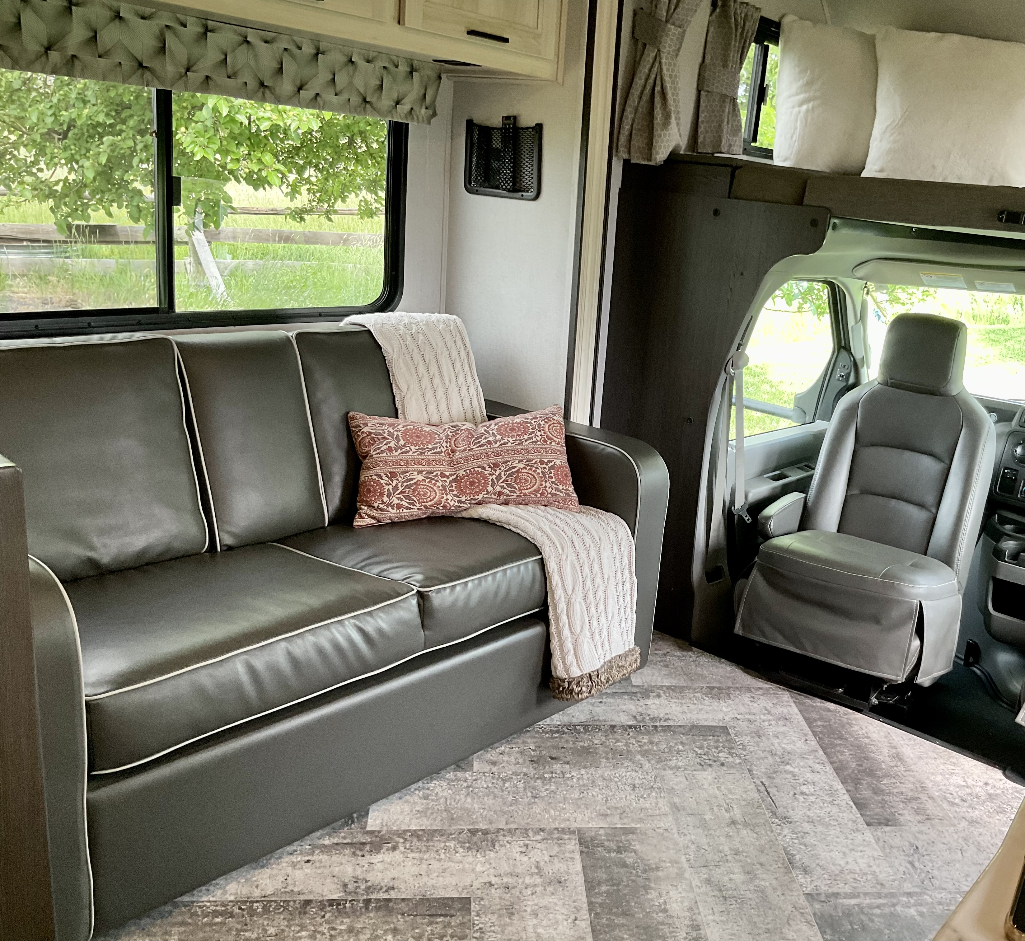 Cozy RV rental interior with a dark sofa, patterned pillow, nature view from window, and bed area above the cockpit.. Forest River Sunseeker 2020