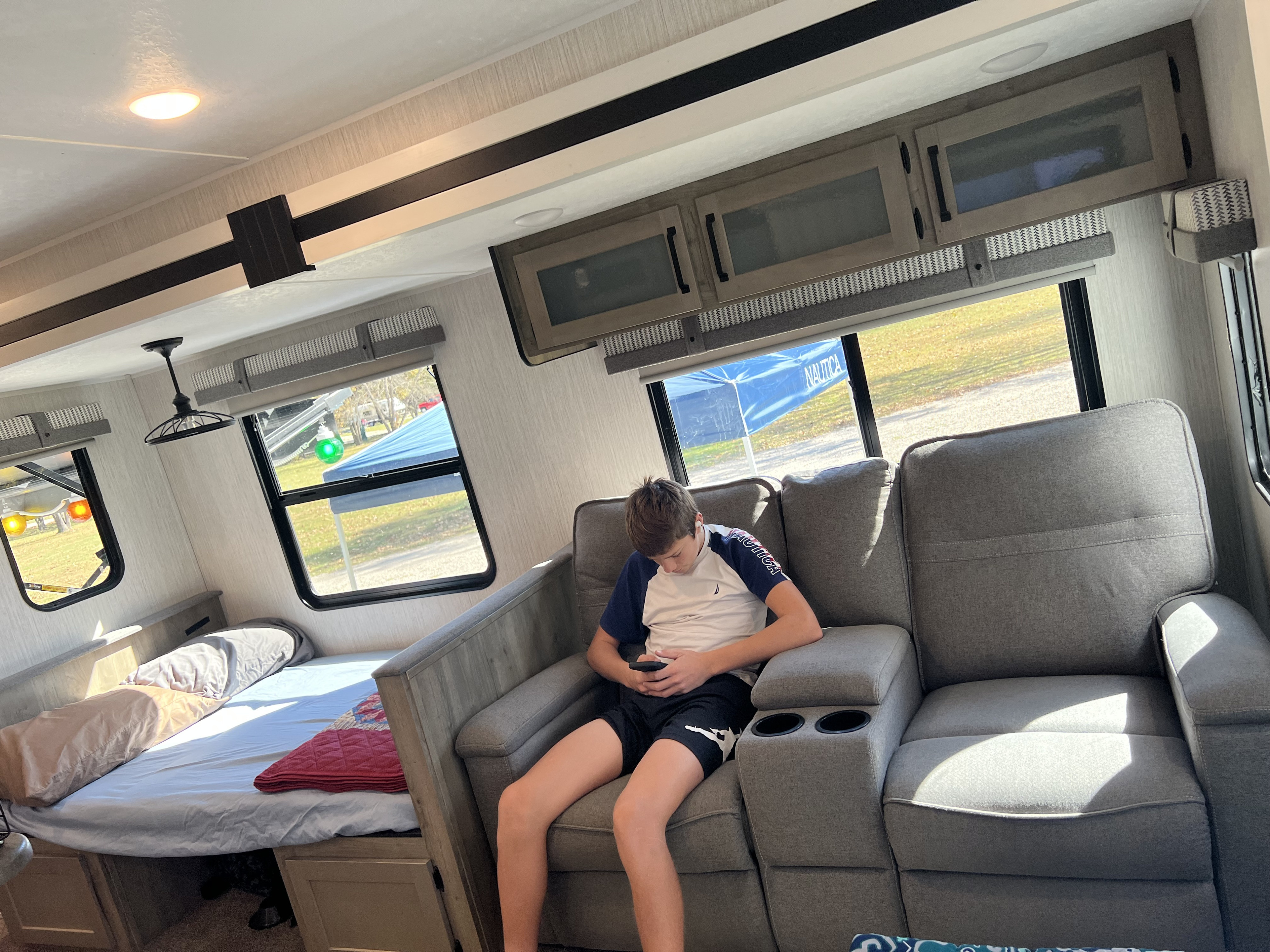 Inside an RV rental: boy on gray sofa with phone. Cozy interior with bed, cabinets, and outdoor views.. Palomino Puma 2021